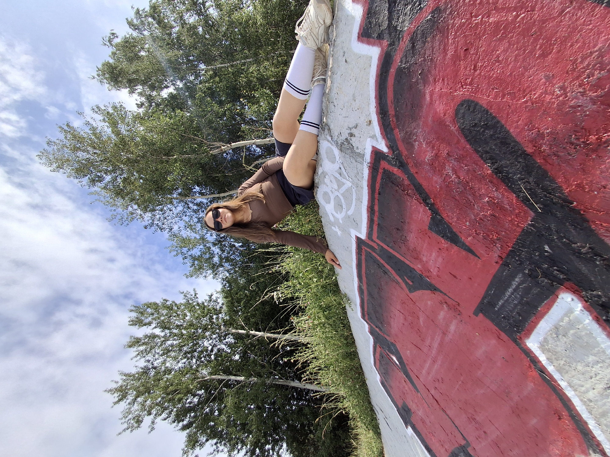A young woman with long brown hair and sunglasses is lying on a concrete surface covered in red and black graffiti. She is wearing a brown long-sleeved shirt, black shorts, white socks with black stripes, and athletic shoes. Her legs are bent, and her right arm is extended, resting on the graffiti. The setting appears to be outdoors, with green trees and a cloudy blue sky visible in the background. The perspective is from a low angle, looking up towards the sky and the woman. The lighting suggests it is daytime. The overall mood appears casual and relaxed.