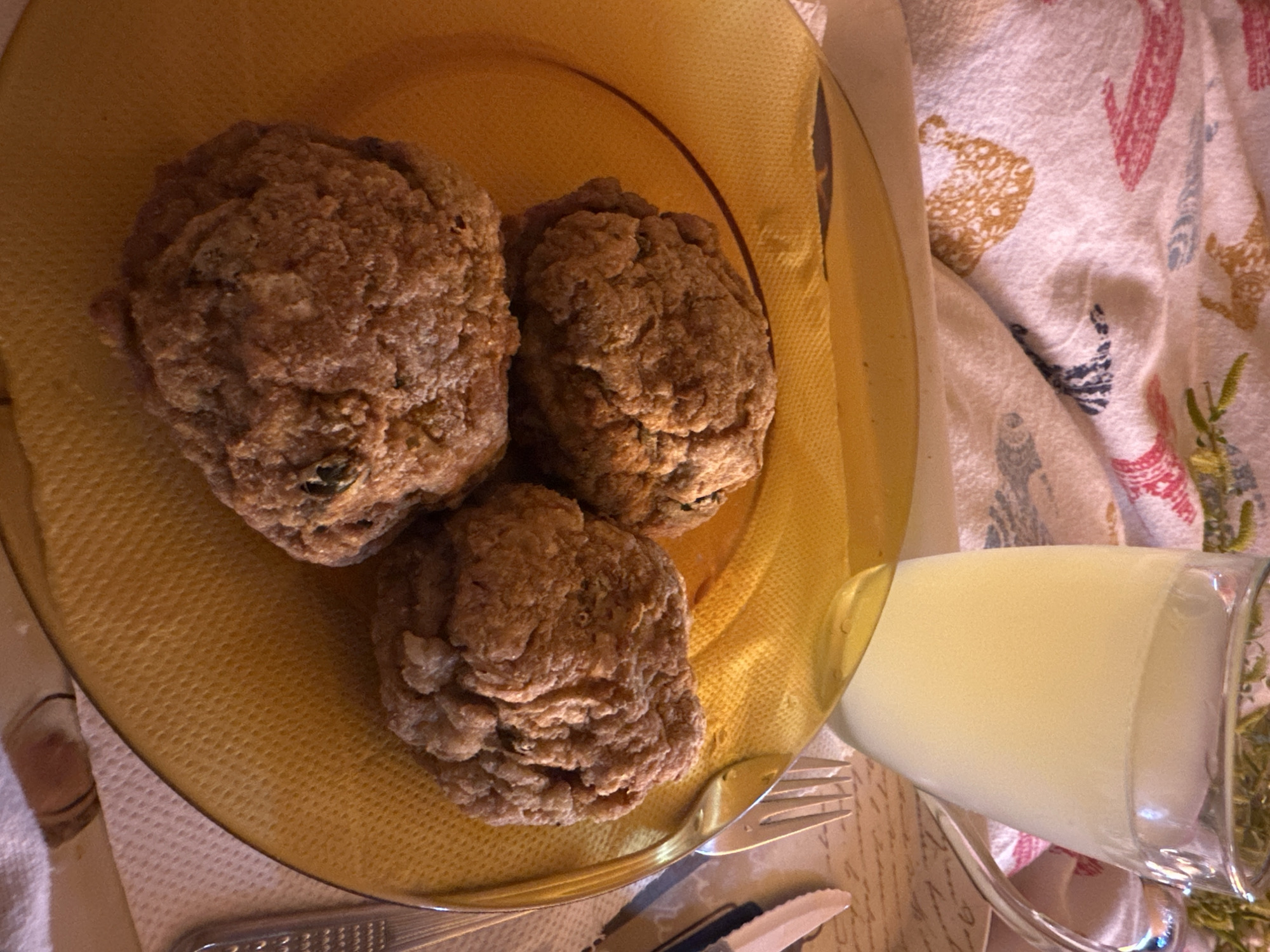 A top-down view shows a yellow plate with three fried patties, a glass of cloudy liquid (likely lemonade), a fork, and a knife on a patterned cloth. The scene is illuminated by warm light, suggesting an indoor setting. There is no indication of the location being Sofia, Bulgaria. There are no people visible in the image.