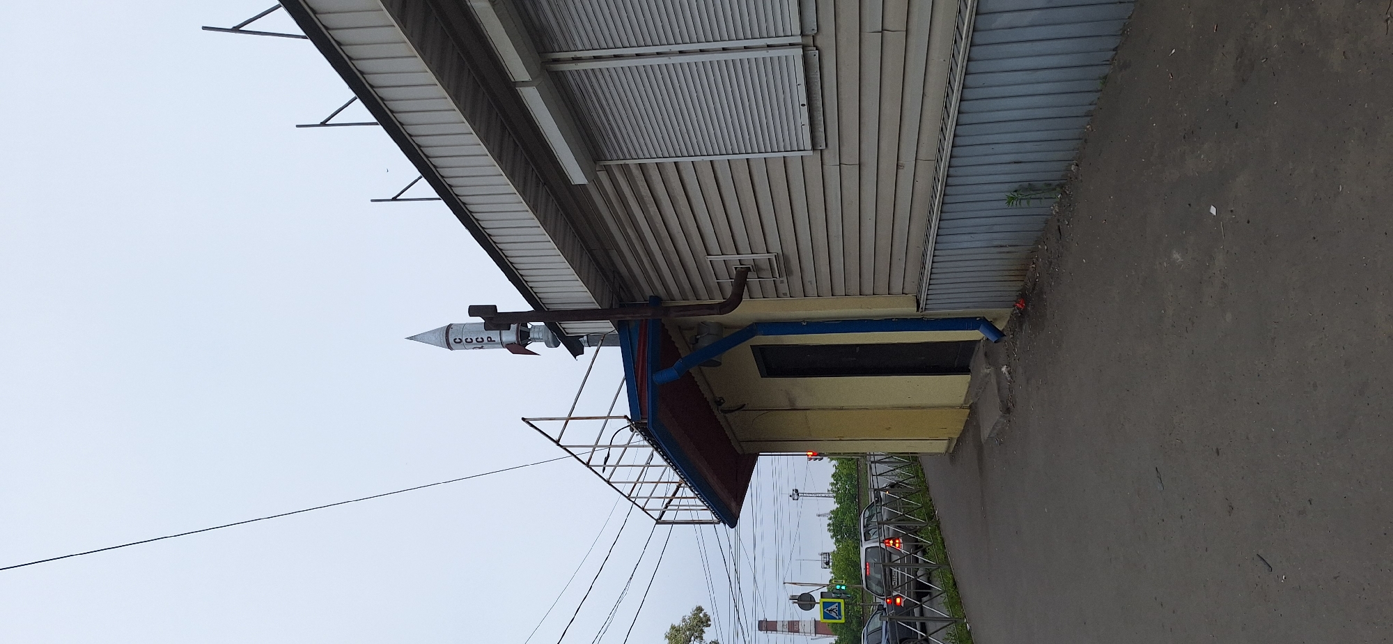 The image captures a street-level view of a building with a rocket-shaped decorative element on its roof. The rocket, painted silver with red fins, has the letters "СССР" vertically inscribed on its side. The building itself appears to be a single-story structure with light-colored horizontal siding and a dark-colored metal base. A dark door is visible on the left side of the building, under a covered awning. A blue downspout runs along the side of the building. The building is situated next to a paved road where several cars are visible. A pedestrian crossing signal with a green light is also present. In the background, industrial structures and trees can be seen under an overcast sky. The overall impression is of an outdoor urban setting.