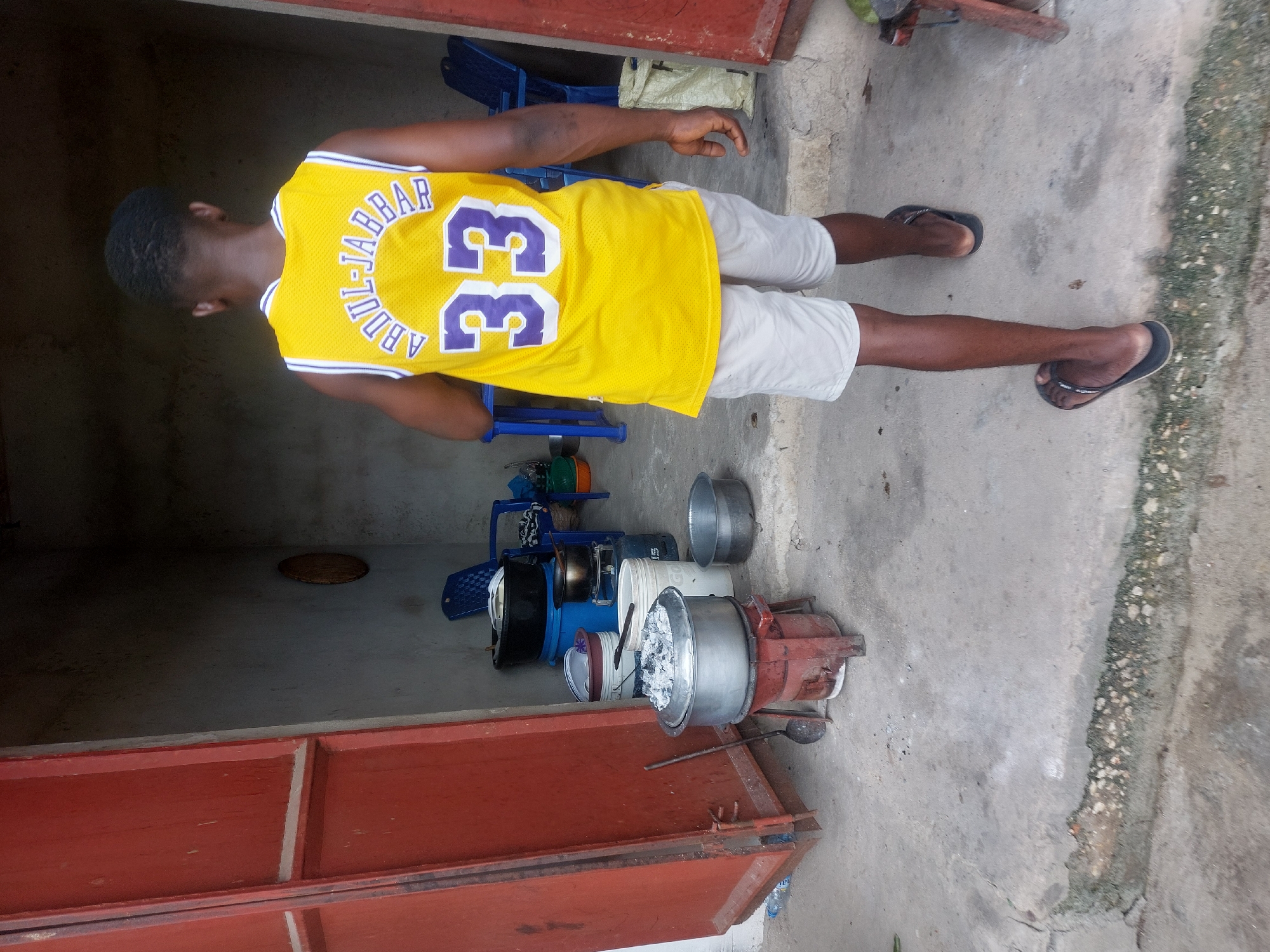 A full-body shot of a person facing away from the camera wearing a yellow basketball jersey with the name "ABDUL-JABBAR" and the number "33" on the back, standing on a concrete surface next to cooking equipment, and wearing light shorts and dark sandals.
The cooking equipment includes large pots, a red cooking stove with hot charcoal on top, metal lids, a white bucket, and cooking utensils.
The location is a concrete area next to a building with a red door.
There is no definitive evidence to confirm the location is in Dar es-Salaam, Tanzania.
