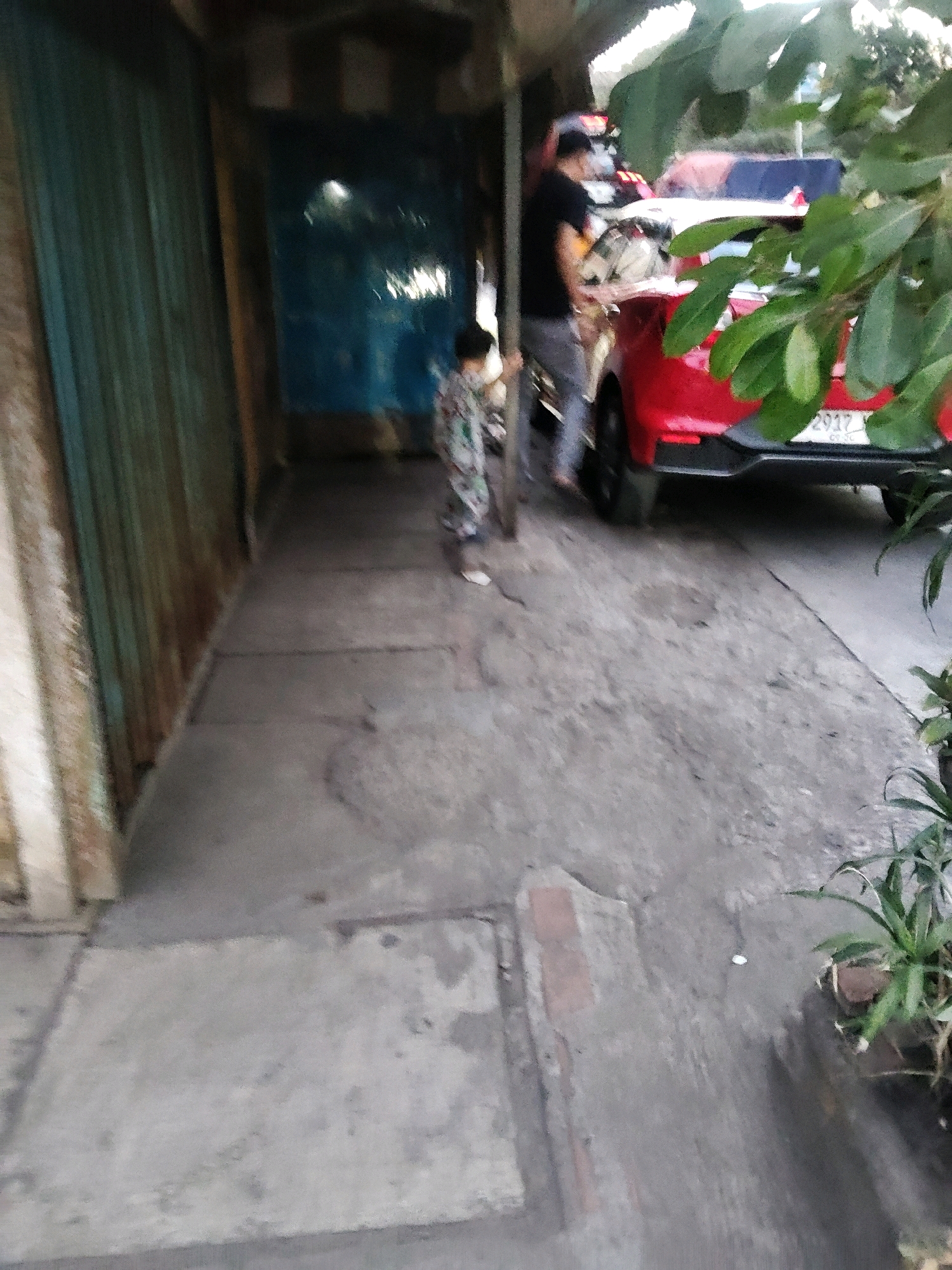 A child wearing a patterned shirt and pants walks along a narrow pathway next to a building. To the right of the pathway, a red car is parked. The car has a license plate with the numbers "2917". A person in a black t-shirt and grey pants stands near the car. The building has corrugated metal walls painted green on the left and a blue door or panel on the right. The ground is paved with concrete. The image appears to be taken outdoors in a built-up area, possibly a street or alleyway in Sukaresmi, Indonesia, given the presence of the car's license plate and the general urban environment.