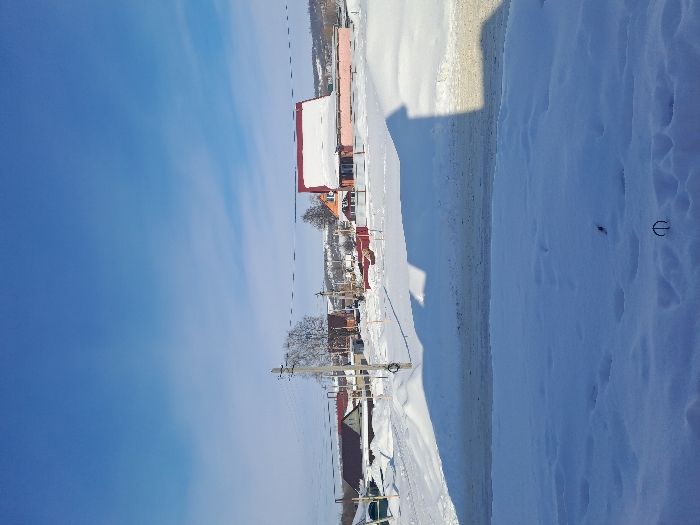 The image displays a snow-covered residential street in Bolshaya Elhovka, Russia, under a clear blue sky with minimal high-altitude clouds. The foreground consists of deep, undisturbed snow, exhibiting a few indistinct indentations and a dark, curved metallic object protruding from the snow near the bottom center. A horizontal strip of dark, partially cleared road separates the immediate foreground from the mid-ground. Long, dark shadows, indicative of structures outside the frame, are cast across the foreground and road, extending from left to right. In the mid-ground, a series of utility poles with multiple overhead wires is visible, along with a bare deciduous tree behind a central pole. Numerous single to two-story residential buildings are present, featuring pitched roofs, many of which are heavily covered in snow. A prominent light pinkish-orange building with a reddish roof and significant snow accumulation is visible on the right side of the frame. The background reveals more scattered structures and distant, snow-dusted hills. No individuals or active subjects are visible in the scene.