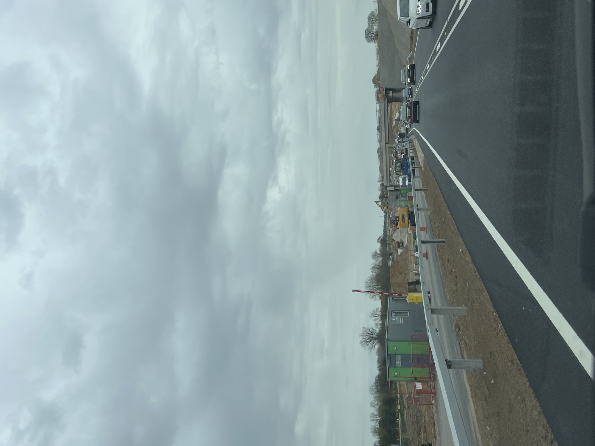 The image shows a road construction site adjacent to a highway. Several vehicles are driving on the highway. Construction equipment, including an excavator, is visible on the construction site. Portable buildings are also present near the construction. The sky is overcast. The location is likely Broadland, United Kingdom, given the context.