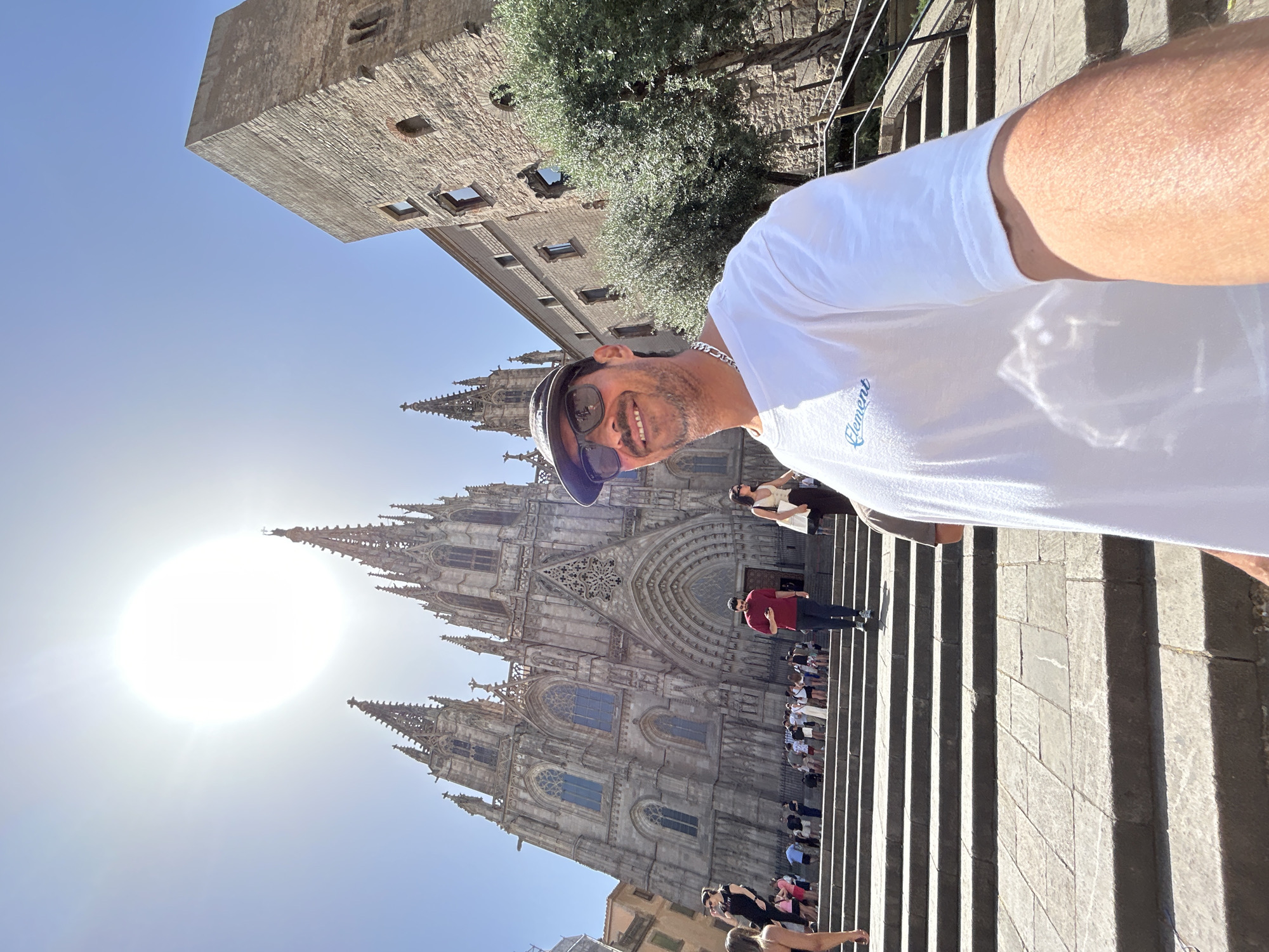 A man wearing a white t-shirt, dark sunglasses, and a baseball cap is taking a selfie in front of the Barcelona Cathedral, located in Barcelona, Spain. The sun is shining brightly in the sky, creating a lens flare. The cathedral is a large Gothic structure with intricate details and tall spires. Many people are visible on the steps leading up to the cathedral, some of whom appear to be tourists. To the right of the cathedral, there is a stone tower and some trees. The man is smiling at the camera.