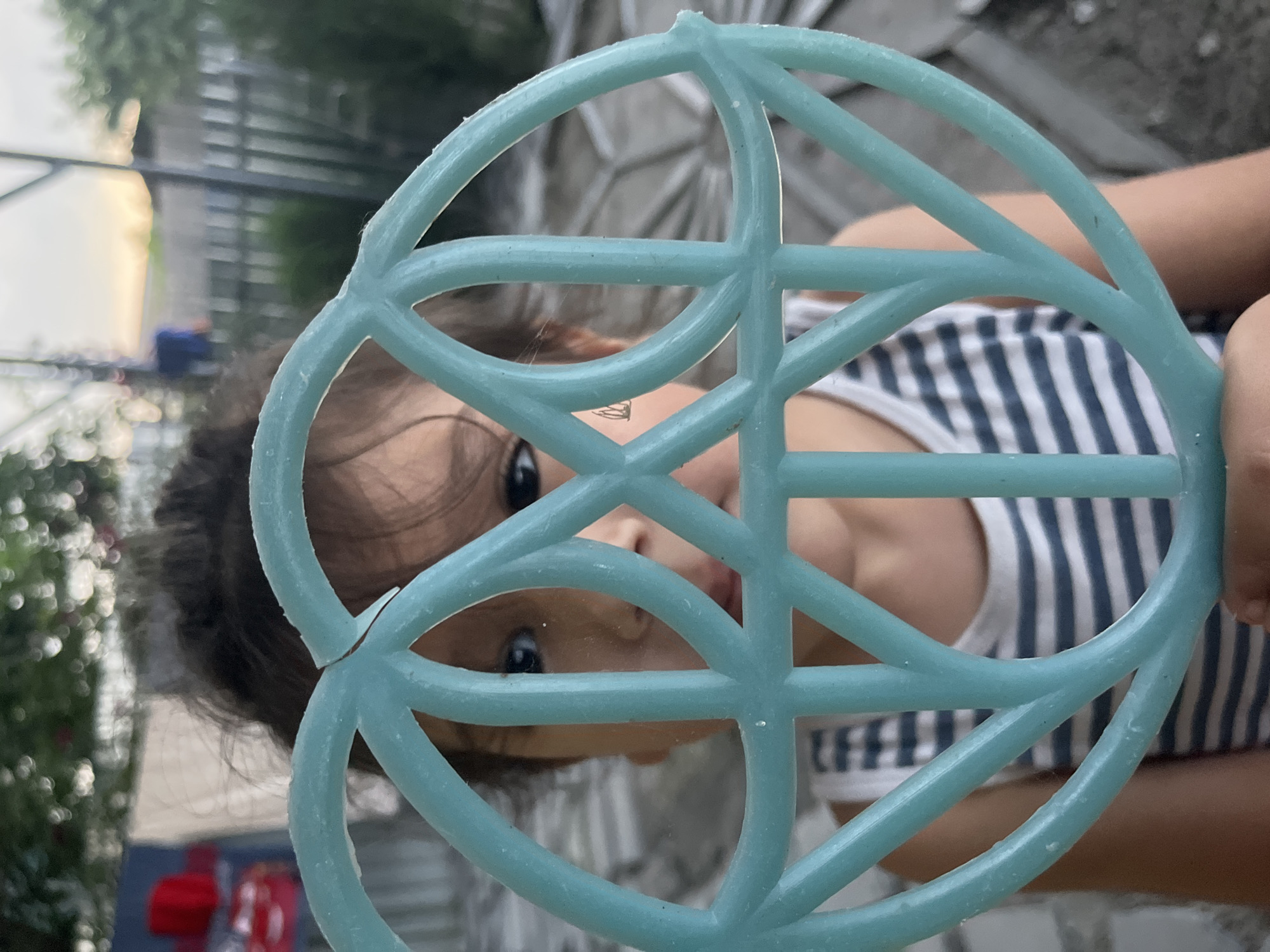 A young child, obscured by a broken teal-colored plastic object, peers through its circular pattern. The child's face is partially visible, with dark hair pulled back and intense eyes. They wear a striped tank top. The background is blurred, suggesting an outdoor setting with some greenery and fencing, possibly a backyard in Tbilisi, Georgia. The overall impression is one of playful curiosity captured in a candid moment.
