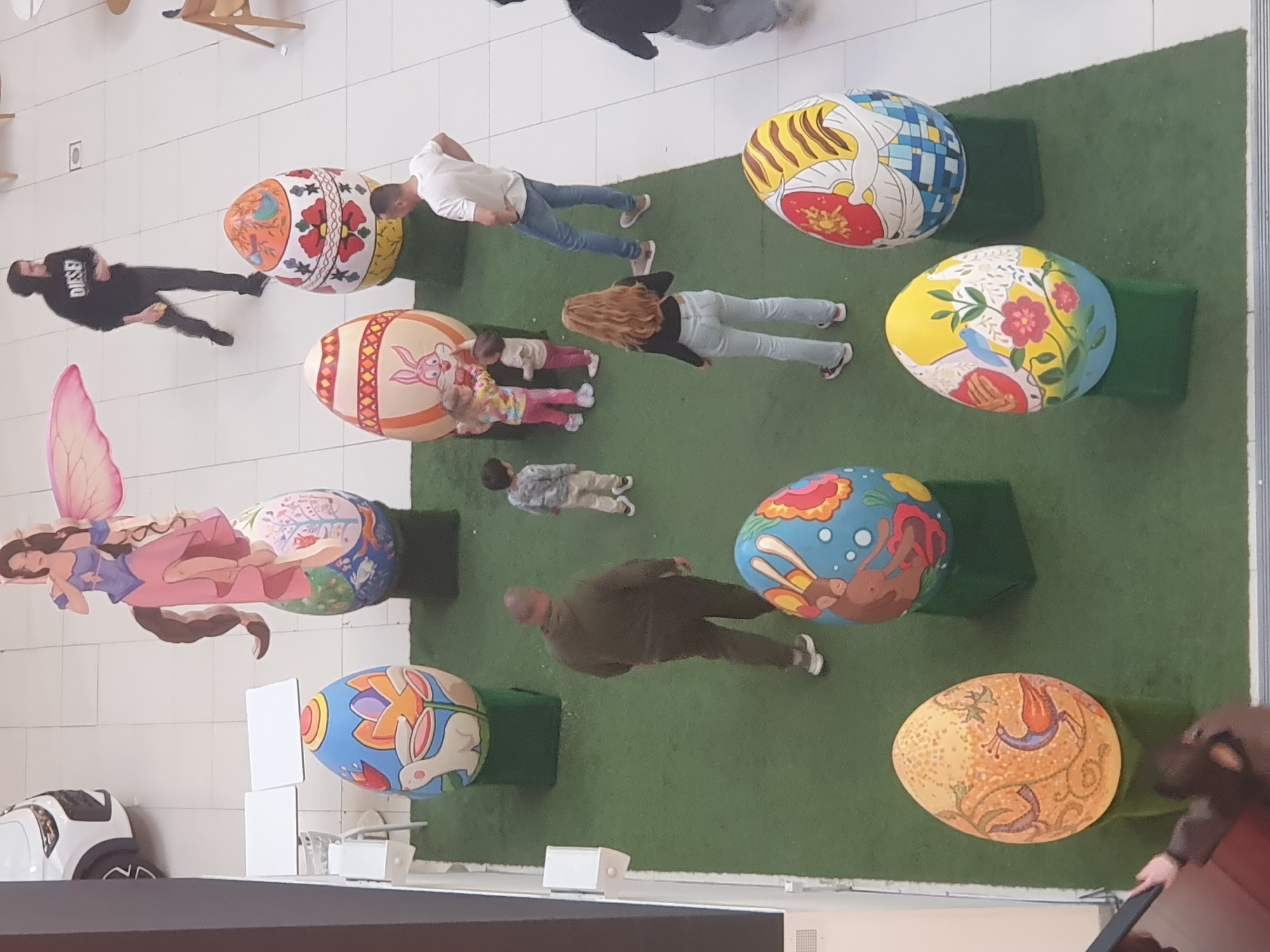 The image depicts a top-down view of an indoor exhibit of large, painted Easter eggs. It seems to be located in a public space, possibly a shopping center or atrium, with tiled flooring on one side and artificial grass on the other.
Several people are present, including a man in a black shirt, a man in a white shirt, a woman with long hair, a man in dark clothing, and several children. The people are scattered around the large Easter eggs, appearing to observe or interact with them.
The Easter eggs are decorated with various designs, including floral patterns, animals (rabbits, birds), and geometric shapes. Some eggs are placed on green, rectangular pedestals, presumably to provide a stable base.
Additional objects include a fairy decoration and what appears to be a toy car. The environment is well-lit, suggesting an indoor location with adequate lighting. The absence of specific landmarks makes it difficult to definitively identify the location within Plovdiv, Bulgaria.