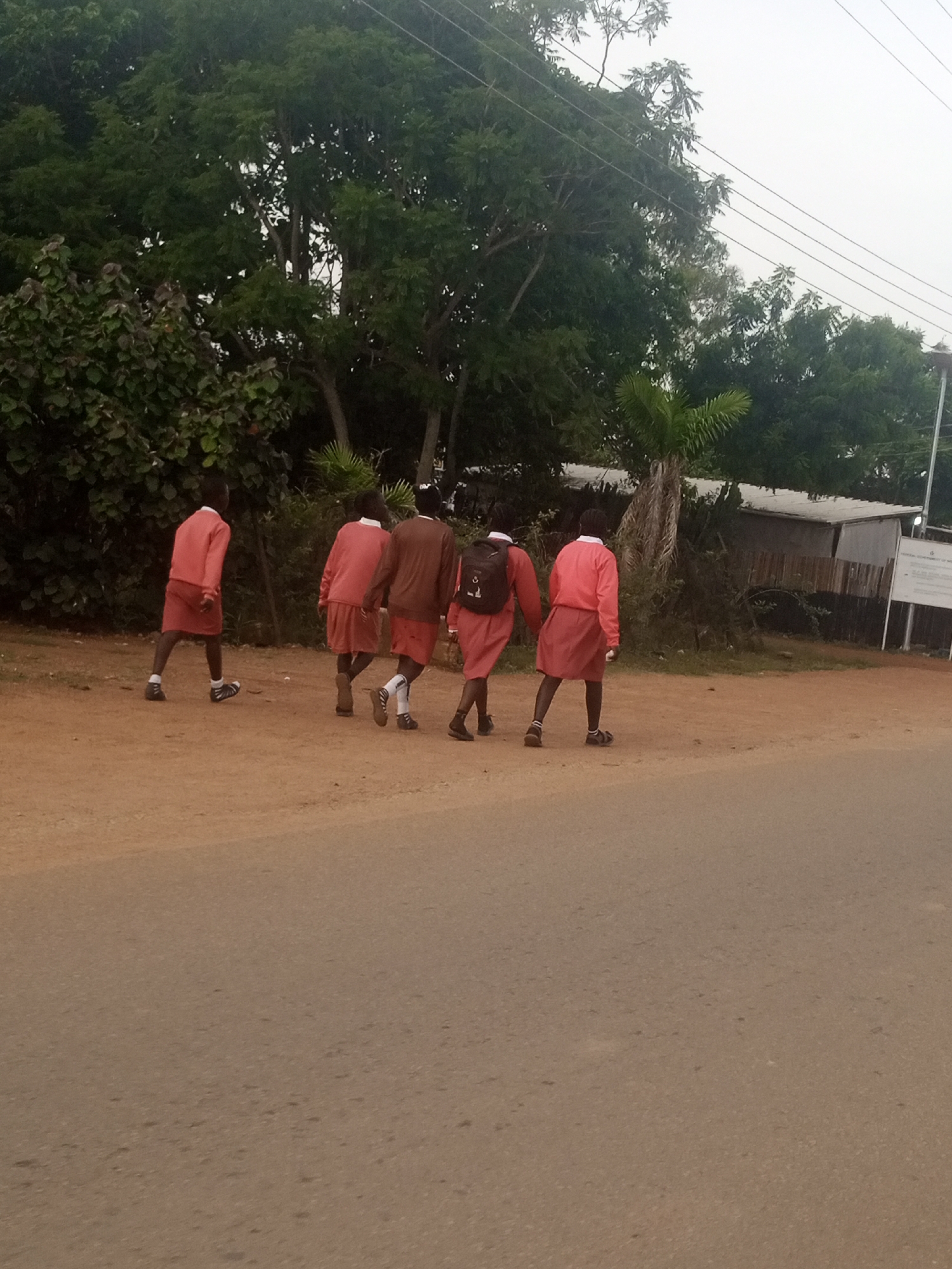 Five young individuals, likely students, are walking on a dirt path next to a paved road. They are all wearing similar uniforms: reddish-pink shirts and skirts, with one individual in a brown top. Three of them have backpacks. The background consists of lush green trees and vegetation, a simple building with a corrugated metal roof, and utility poles with wires. The sky is overcast. The scene appears to be in a rural or semi-urban area.