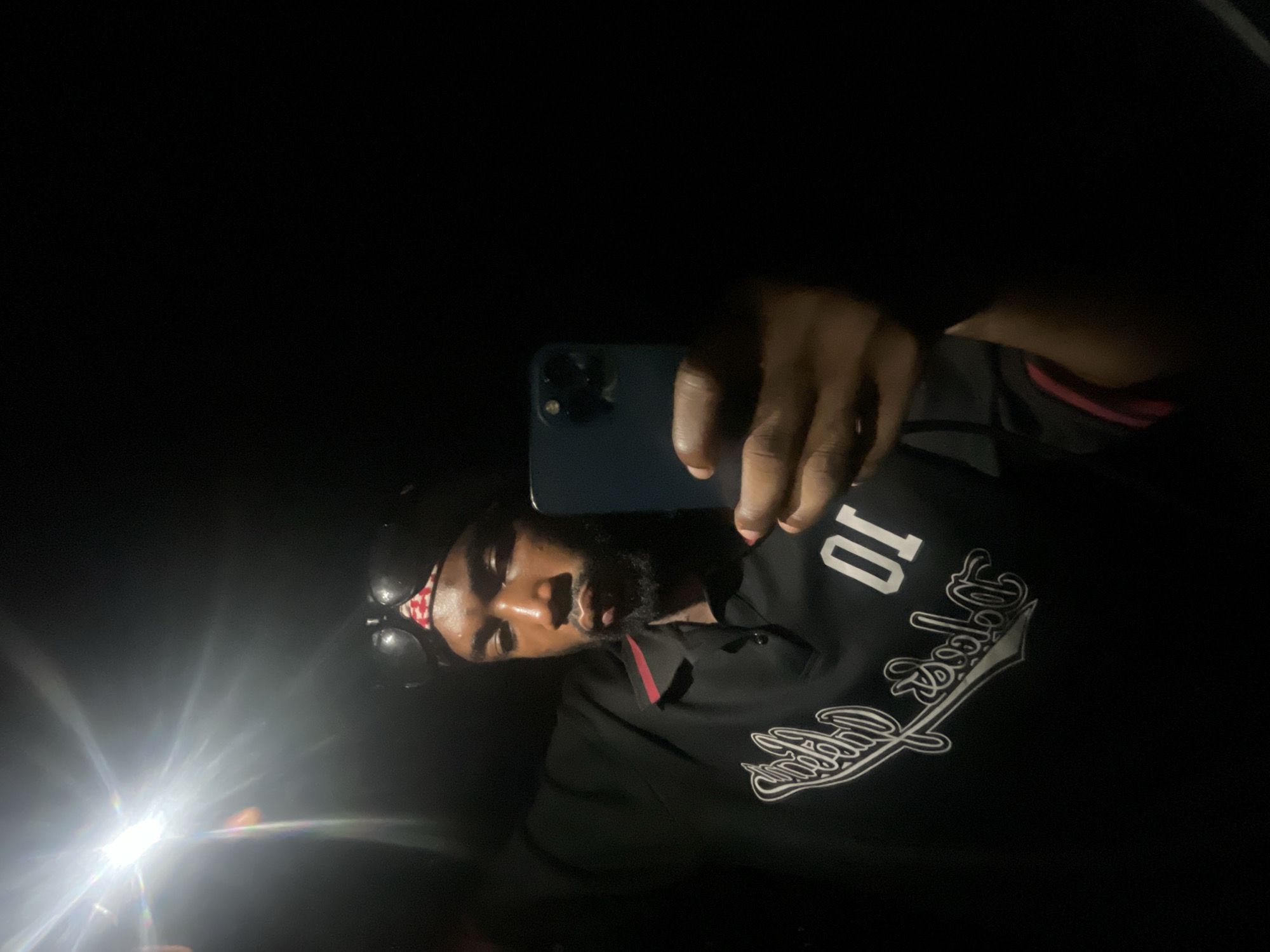 A person is taking a selfie in a dark environment, possibly at night. The person is wearing a black shirt with a white design and the number "10" on it. They are also wearing a red and white patterned bandana and sunglasses pushed up on their head. The person is holding a blue smartphone, using its flashlight as a light source. The location is unknown and cannot be determined as Lokoja, Nigeria.