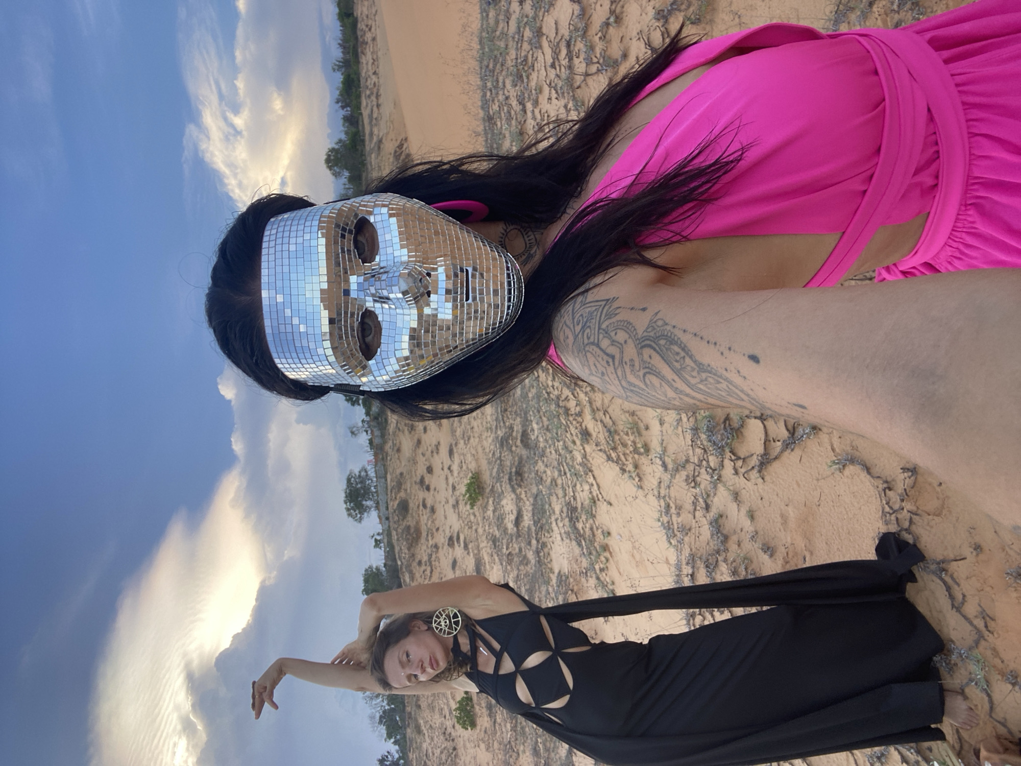 The image depicts two individuals in a desert-like environment, likely Mũi Né, Vietnam. One person in the foreground is wearing a striking, bright pink dress and a silver disco ball mask, obscuring their face. Their long, dark hair is visible, and tattoos adorn their arm. To the left of the mask, glimpses of a bright blue sky with scattered clouds suggest it might be daytime, possibly late afternoon or early evening, given the quality of the light.
In the background, a second person is positioned lower in the frame, wearing an elaborate black dress with geometric cutouts. They are striking a dynamic pose with their arms extended upwards. The setting is characterized by sand dunes with sparse, dry vegetation, characteristic of a coastal desert. Trees can be seen in the distance, suggesting the proximity of a more vegetated area or coastline. The overall mood appears to be artistic and perhaps performative, with the dramatic outfits and the unusual mask. The lighting is soft, indicating a time when the sun is not directly overhead. No visible text is present in the image.