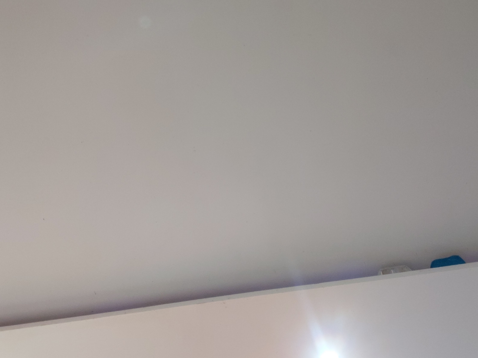 The image presents an upward view of an indoor setting, dominated by a plain, light-colored ceiling that occupies most of the frame. The ceiling appears to be a uniform, possibly off-white or light grey surface, with a few faint specks or imperfections visible upon closer inspection.
At the very bottom of the image, a horizontal line marks the top edge of a white wall or piece of furniture, such as a cabinet or shelf. Resting on this edge, in the bottom right corner, are two small objects. One is a distinctly blue object, which appears soft or rubbery, partially obscured by the edge. To its left, another smaller, light-colored, somewhat translucent object, possibly made of plastic, is visible.
A prominent bright lens flare emanates from the bottom right corner, suggesting a strong light source, possibly artificial, is present just outside the frame. There is no discernible activity taking place, and no people are visible. No text is present in the image. The time of day cannot be determined from the lighting, and there are no specific location cues within the image itself to indicate Santa Ana, Venezuela.