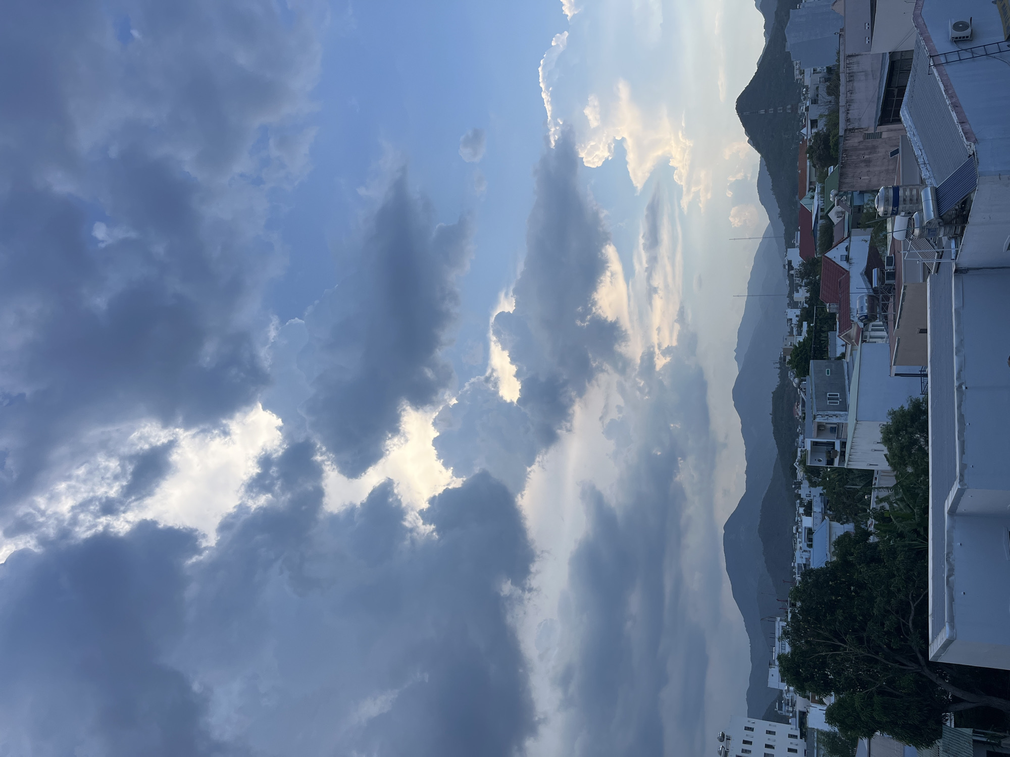 The image shows a view of Bac Nha Trang Ward, Vietnam. The foreground is dominated by a sky filled with dark, heavy cumulonimbus clouds. Patches of bright, diffuse sunlight break through the clouds. The midground features a cityscape of low-rise buildings with a mix of roof types. Mountains are visible in the background. There are trees interspersed among the buildings. The image appears to be taken from an elevated vantage point, like a rooftop. No people are visible.