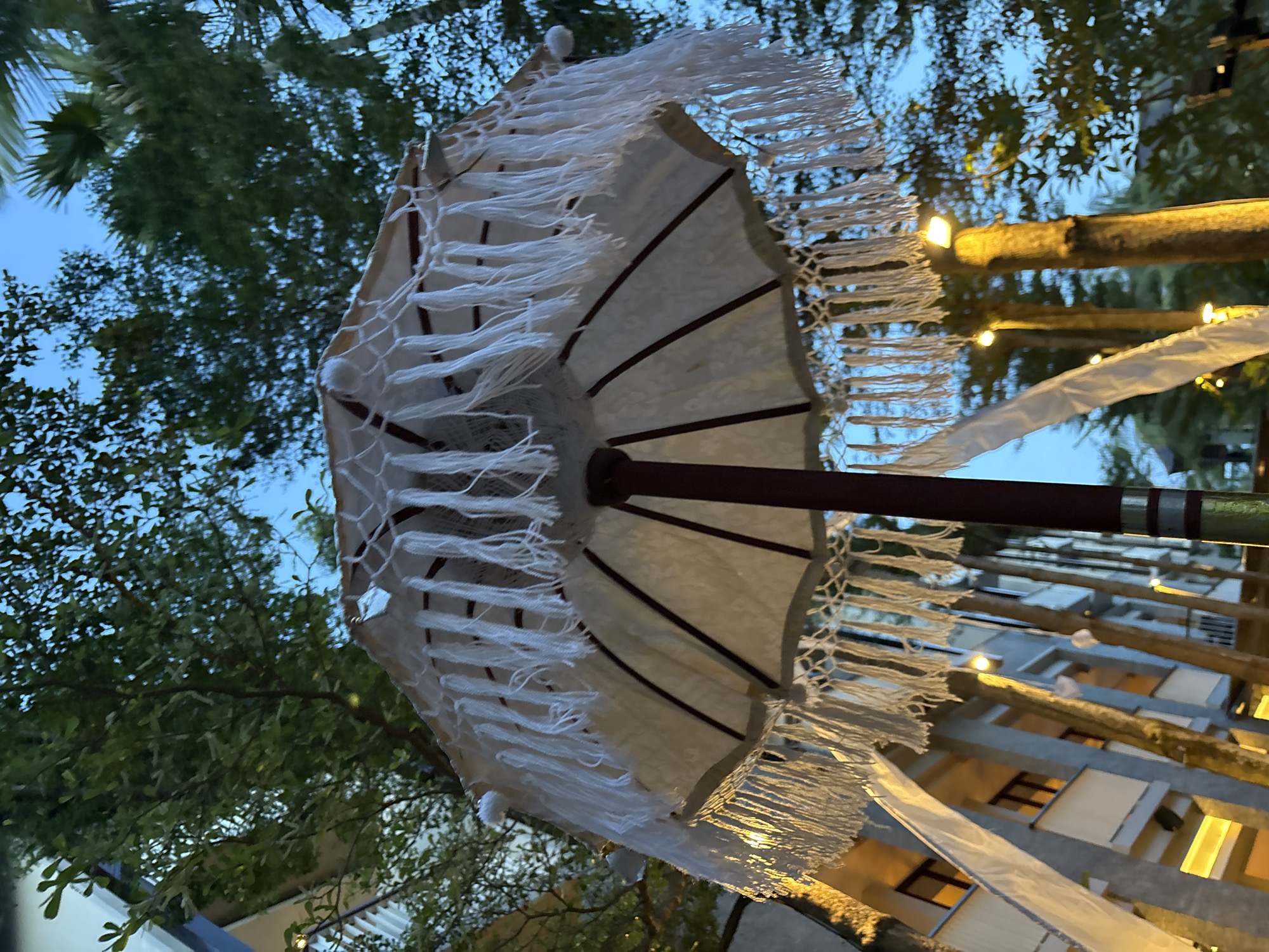 The image depicts a decorated umbrella in Kuta Selatan, Indonesia. The umbrella is white with tassels around the edge and appears to be part of an outdoor setting, possibly for an event or decorative purpose. The umbrella is opened, and the structure is partially visible. The umbrella is in the vicinity of green trees and buildings, with some string lights visible on the side. The overall scene suggests an outdoor setting, possibly a patio, park, or street, with decorations. The lighting seems to indicate it is either evening or early morning.