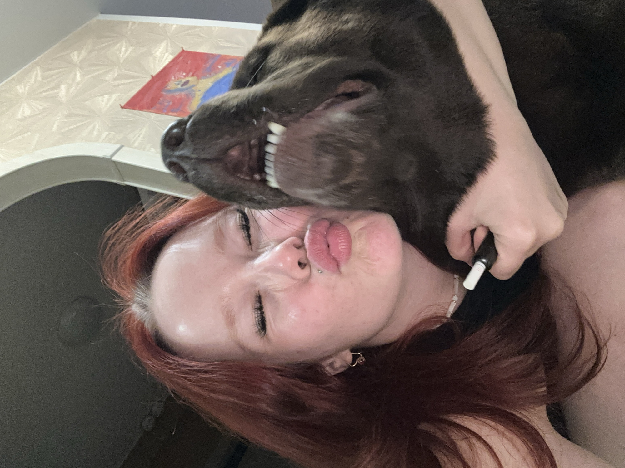 A young woman with auburn hair is taking a selfie with a dark-colored dog. The woman is puckering her lips and closing her eyes, while the dog is sticking its head towards the camera, showing its teeth. The background is out of focus but appears to be an indoor setting with patterned wallpaper. The woman is wearing a black top and a beaded necklace. The dog's snout and part of its head are visible.