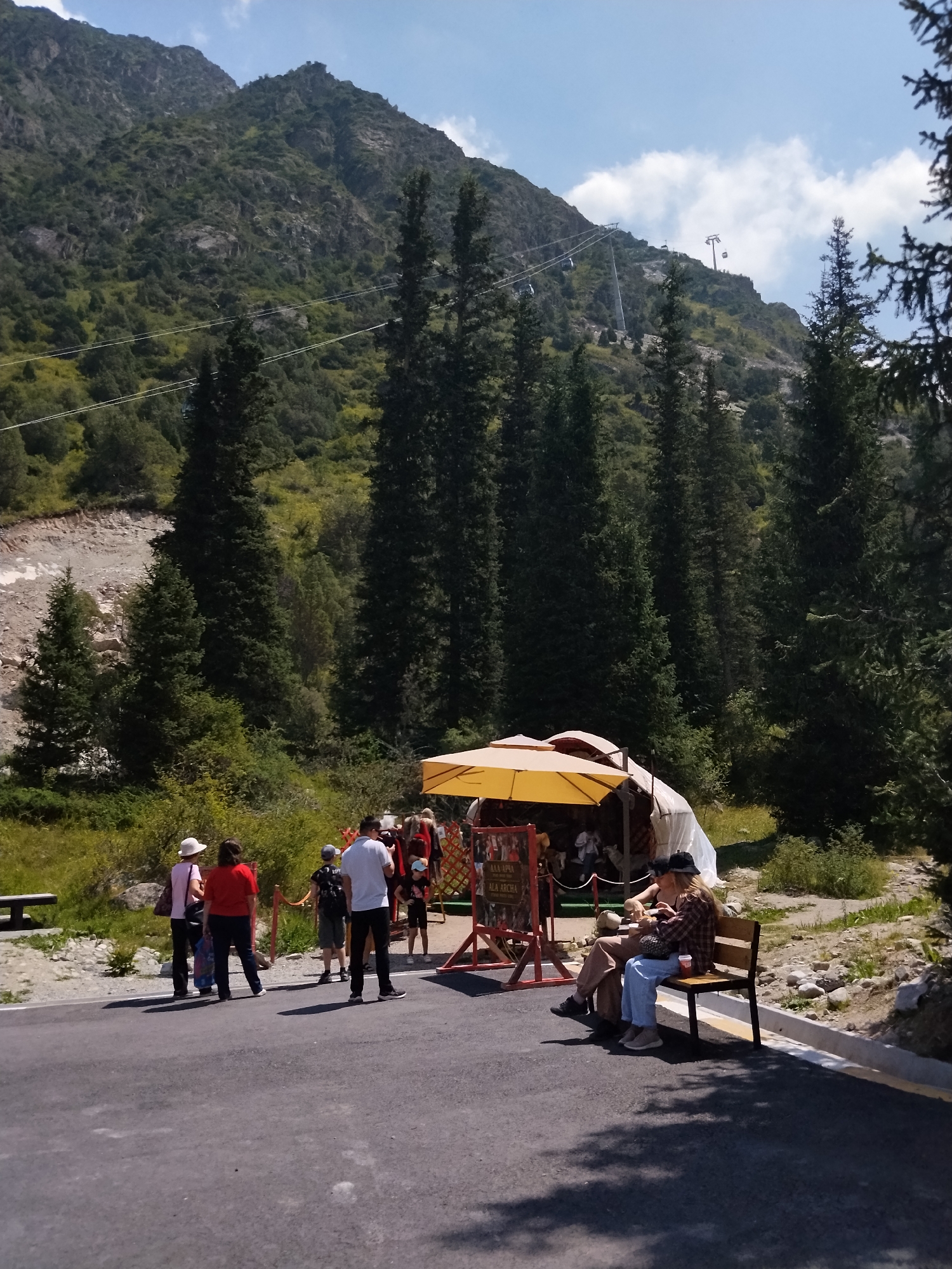 In the mountainous terrain of an unknown location in Kyrgyzstan, a group of people are gathered near a yellow-canopied structure, possibly a vendor stall or rest area. Several individuals stand in a line, facing the stall, while two people are seated on a wooden bench to the right. One person on the bench is wearing a plaid shirt and blue jeans, while the other is wearing a brown jacket and light-colored pants. The environment is characterized by steep, green, and rocky mountainsides, dotted with numerous tall evergreen trees. A cable car system is visible ascending the mountains in the background. The ground is paved in the foreground, with a gravelly path and vegetation to the sides. The sky is clear with a few scattered white clouds.