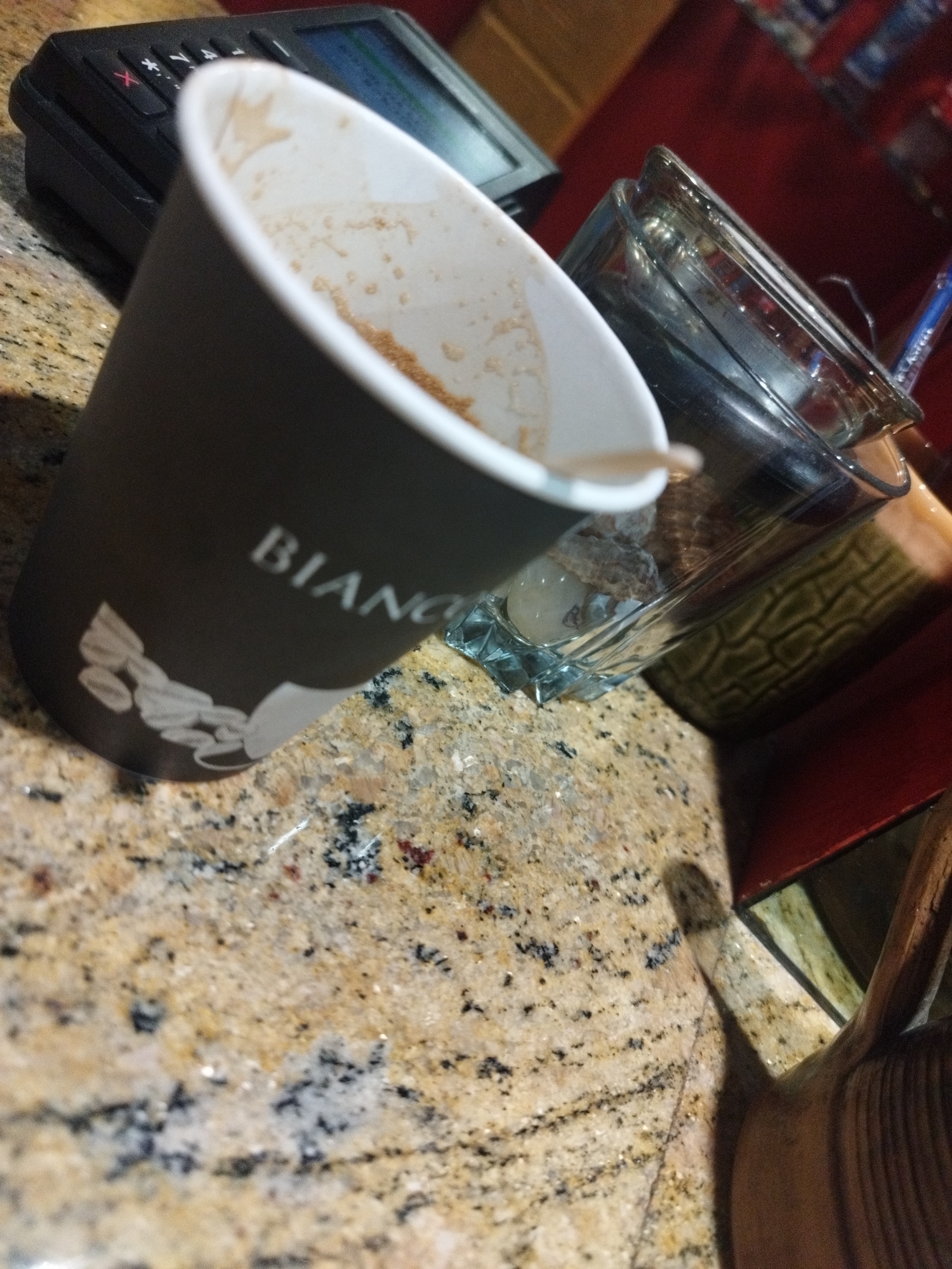 The image depicts a close-up, angled view of a brown disposable coffee cup with the word "BIANCO" and a graphic of coffee beans printed on it, resting on a speckled granite countertop. The cup contains a small amount of coffee residue. To the right of the cup is a clear glass jar with a patterned object inside and a blue pen leaning against it. In the background, a black credit card terminal is visible. The overall setting suggests a cafe or similar establishment. The image does not provide any information about the city of Smolyan, Bulgaria, or any specific actions or interactions.