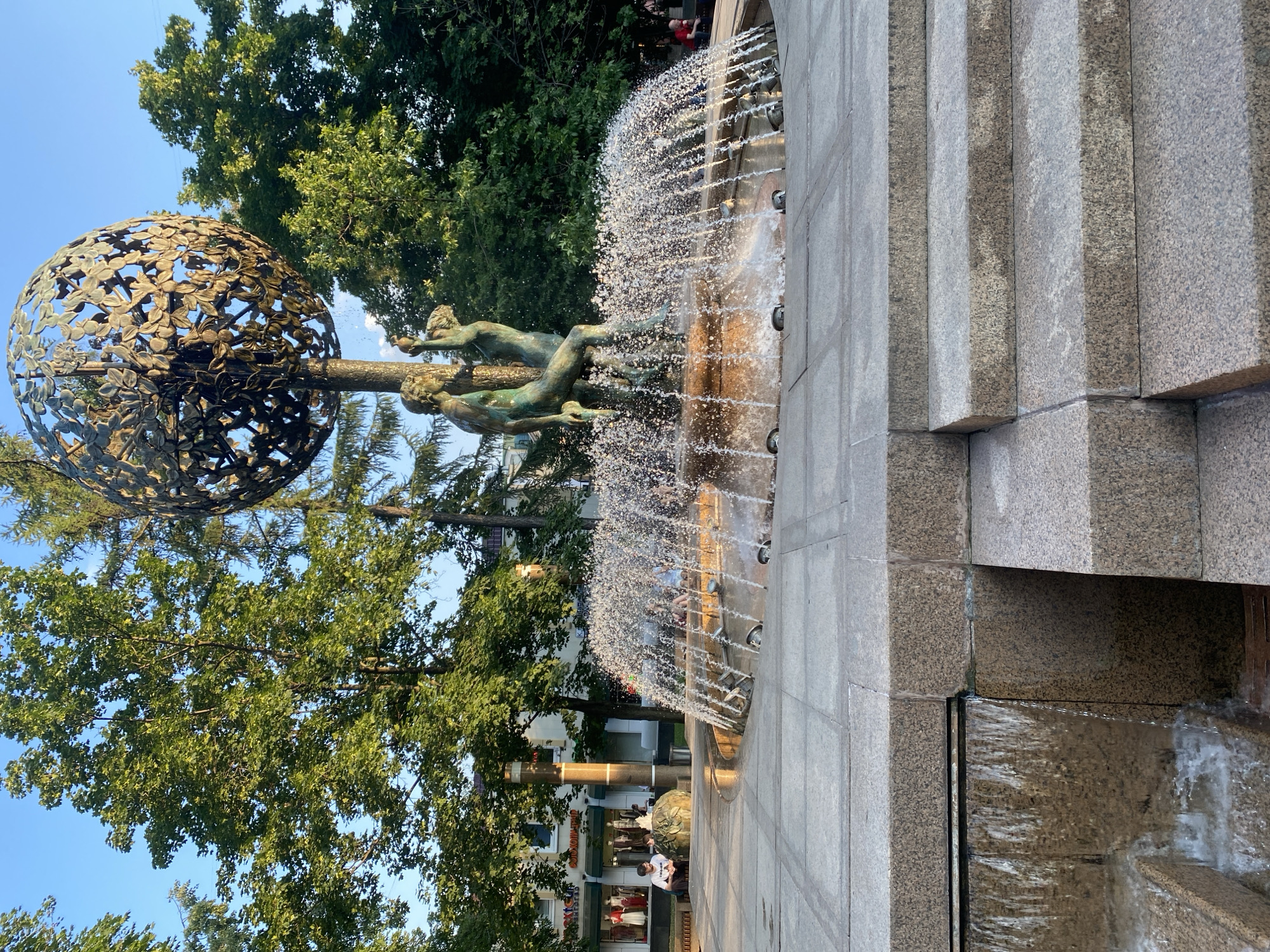 The image depicts a dynamic outdoor scene centered around a decorative fountain. The main subject is a large, ornate sphere made of metal, resembling a globe covered in intricate leaf patterns. Emerging from the globe is a structure supporting several bronze figures of people, some appearing to interact with the water. The fountain is active, with water spraying outwards, creating a misty effect that catches the light.
The fountain is situated on a tiered structure made of granite-like stone, with distinct rectangular blocks forming the steps and edges. Water cascades down these steps, adding to the visual and auditory experience of the fountain. Lush green trees with dense foliage frame the scene, suggesting a park-like or urban green space. The sky above is a clear, bright blue, indicating fair weather and likely daytime.
In the background, buildings with multiple stories are visible, some with awnings and shops at street level. People are present in the scene, some walking in the distance, others near the fountain's edge, enjoying the public space. The lighting suggests it is late afternoon or early evening, with the sun casting a warm glow on the water and stone. The overall mood is one of a lively, public gathering space on a pleasant day. There is no visible text in the image.
