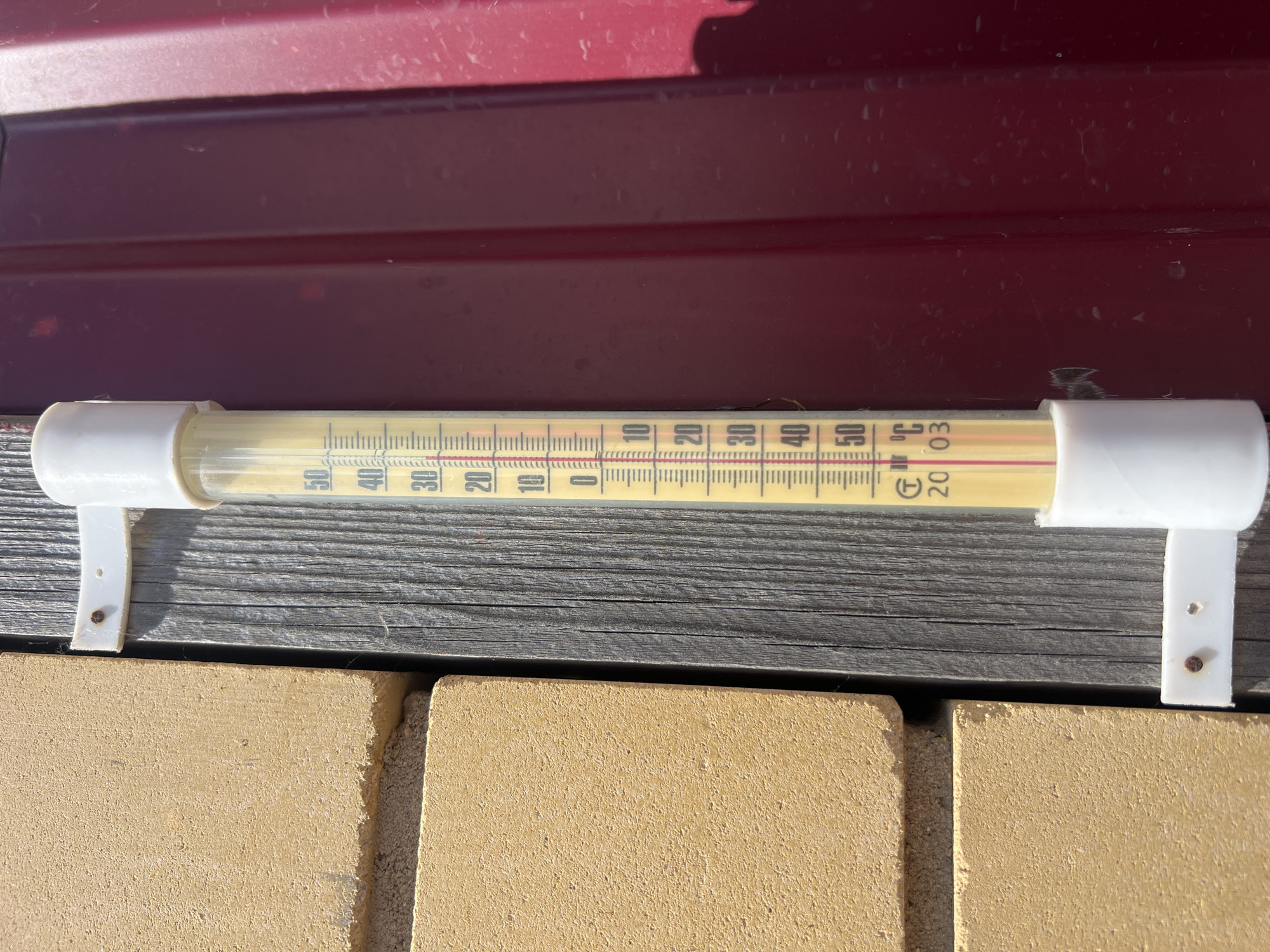 The image shows a thermometer mounted on a wooden surface. The thermometer is a glass tube filled with a yellow liquid, with markings indicating temperature in Celsius. The markings range from 0 to 50 degrees Celsius. The red liquid level is between the 30 and 40 degree marks, indicating a temperature of approximately 34 degrees Celsius. The thermometer is attached to the wooden surface by two white plastic brackets. The wooden surface appears weathered and is mounted against a red metal surface. Below the wooden surface, there are two yellow-colored bricks. The lighting suggests it is daytime, with sunlight casting shadows. The setting appears to be outdoors, possibly in a garden or on a patio. There is no visible text in the image, other than the temperature markings on the thermometer.
