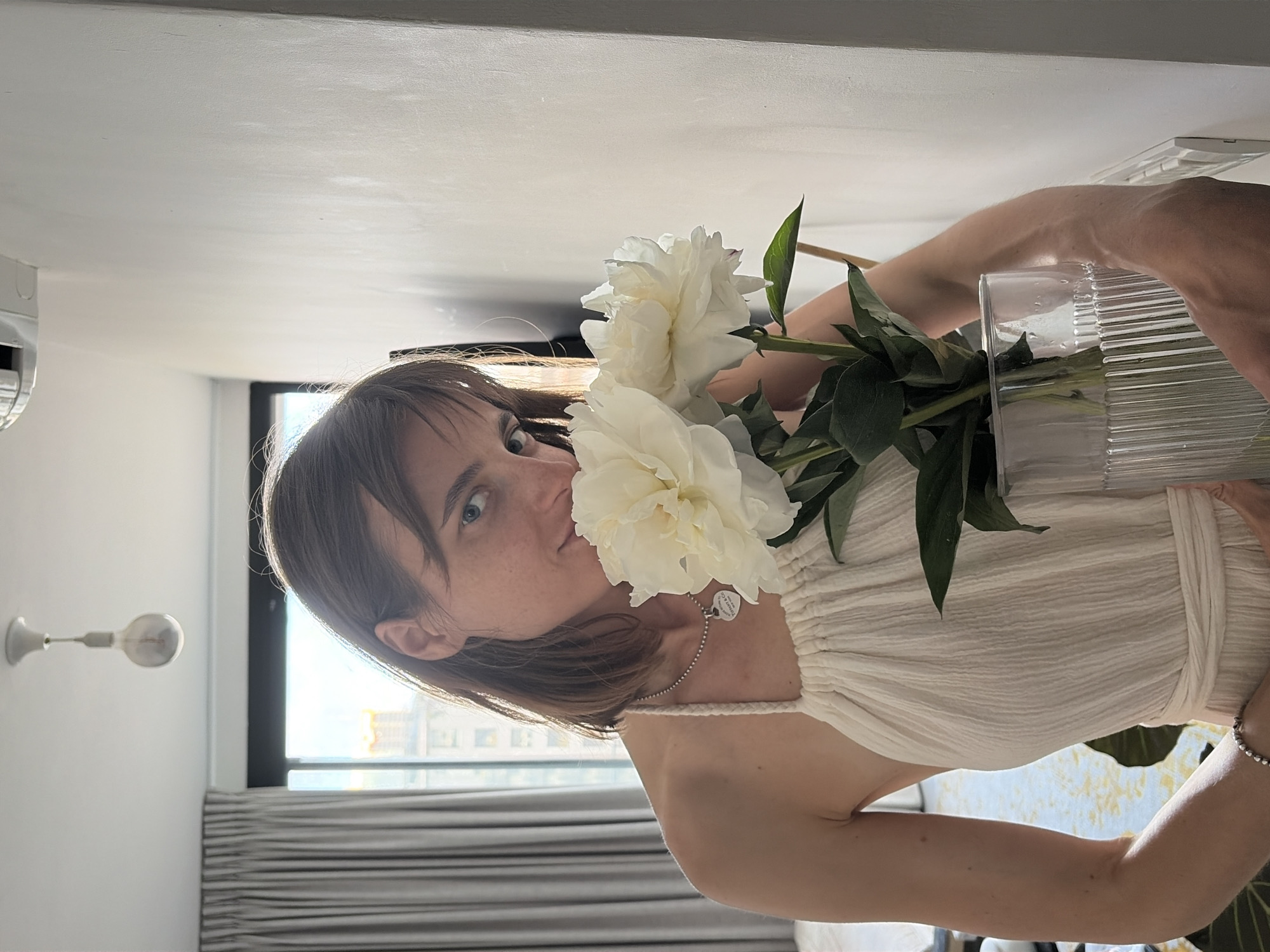 A woman with medium-length brown hair, her face partially obscured by two large white peonies, holds the flowers in a clear glass vase. She is wearing a cream-colored halterneck dress and a silver necklace with a heart-shaped pendant. Her right arm is visible, holding the vase, and her left arm rests across her torso. The background suggests an indoor setting, possibly an apartment, with a large window with grey curtains on the left, revealing a view of buildings and the sky. A light fixture is visible on the ceiling. The overall lighting suggests natural light coming from the window, creating soft shadows. The scene appears to be in Tel-Aviv, Israel, given the architecture visible through the window.