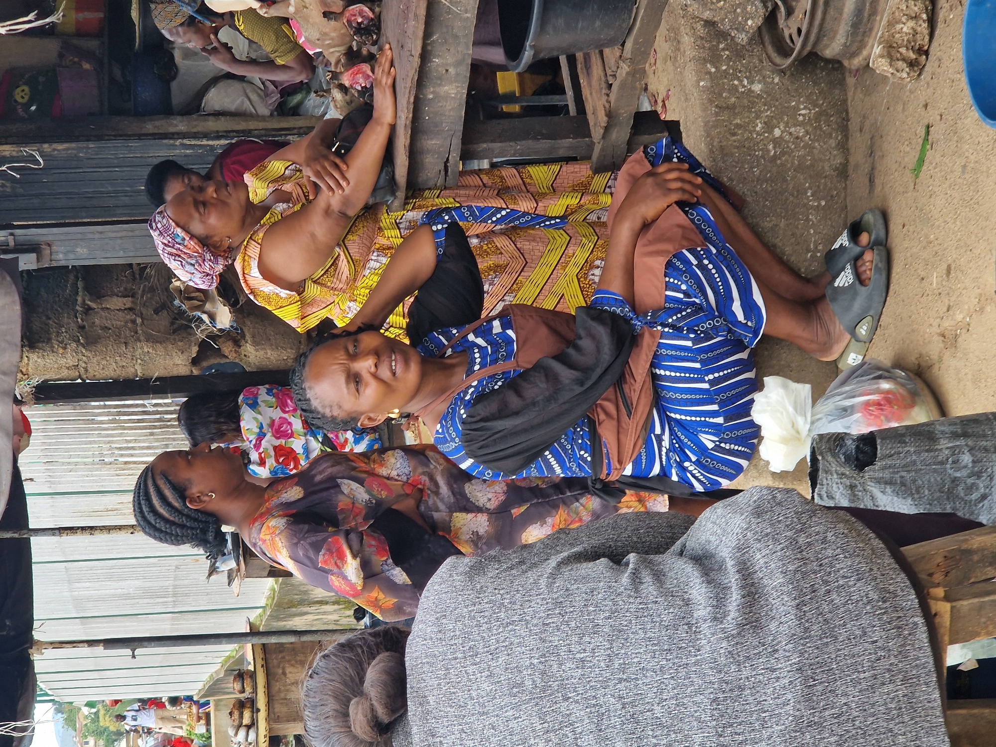 This image depicts a street market scene in Kabong, Nigeria. In the foreground, a woman wearing a blue and white patterned dress and a brown apron is kneeling on a concrete step. Her left hand is resting on her lap, and her right hand is touching her head. She is looking towards the camera with a neutral expression. To her left, a person wearing a grey textured garment is partially visible, with their back to the camera and their hair styled in a bun. Behind the kneeling woman, another woman wearing a yellow and patterned dress with a headscarf is visible. She is leaning on a wooden table, with her arms resting on it. In front of her, on the table, are several pieces of raw meat, possibly goat or sheep, with visible bone and flesh. To the far left of the frame, another woman in a colorful patterned dress is standing, her face in profile, looking to the left. The background shows market stalls with corrugated metal sheeting and wooden structures, suggesting a bustling marketplace. Bags of goods and a large black bucket are also visible. The overall lighting is overcast, suggesting it is daytime.