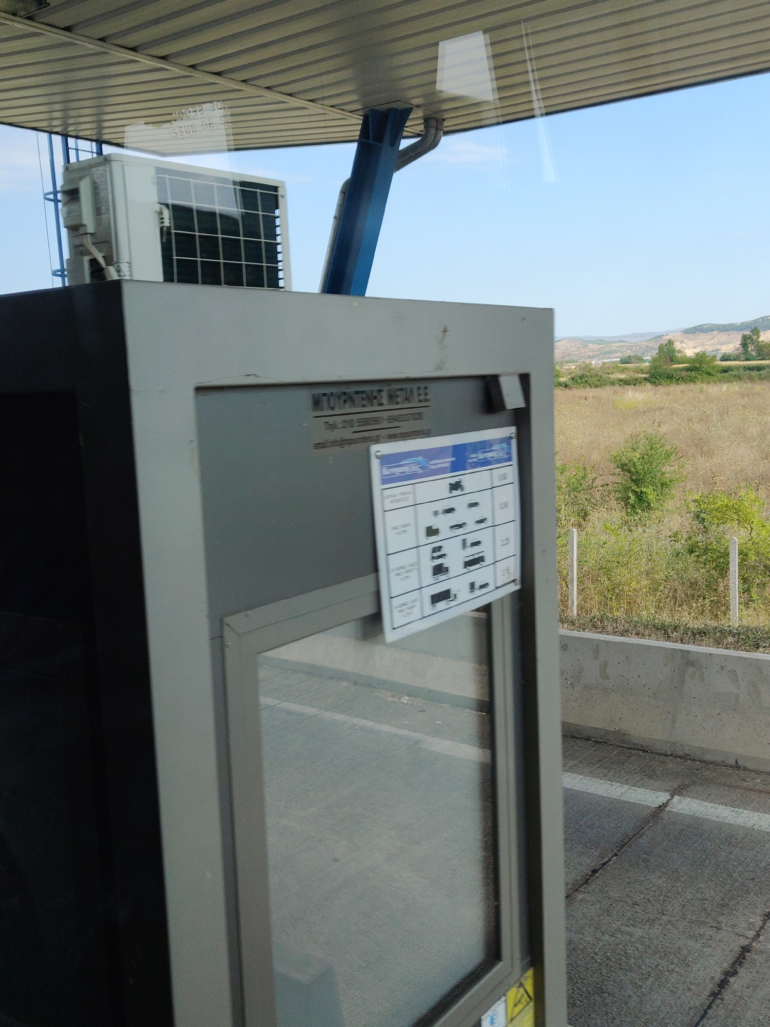 The image shows an exterior shot of a toll booth or service station on a sunny day in Trikala, Greece. The foreground is dominated by a gray, rectangular structure, likely part of the toll booth, with a glass window on the right side. Attached to this structure is a sign with a chart displaying different vehicle types and corresponding charges, presented in Greek. Above the structure, an outdoor air conditioning unit is visible, mounted on a gray base. The background features a dry, grassy field with scattered green bushes and trees, leading to rolling hills under a clear blue sky. A concrete barrier runs along the right side of the image, bordering the road or paved area in front of the toll booth. The overall scene suggests a semi-urban or roadside environment.