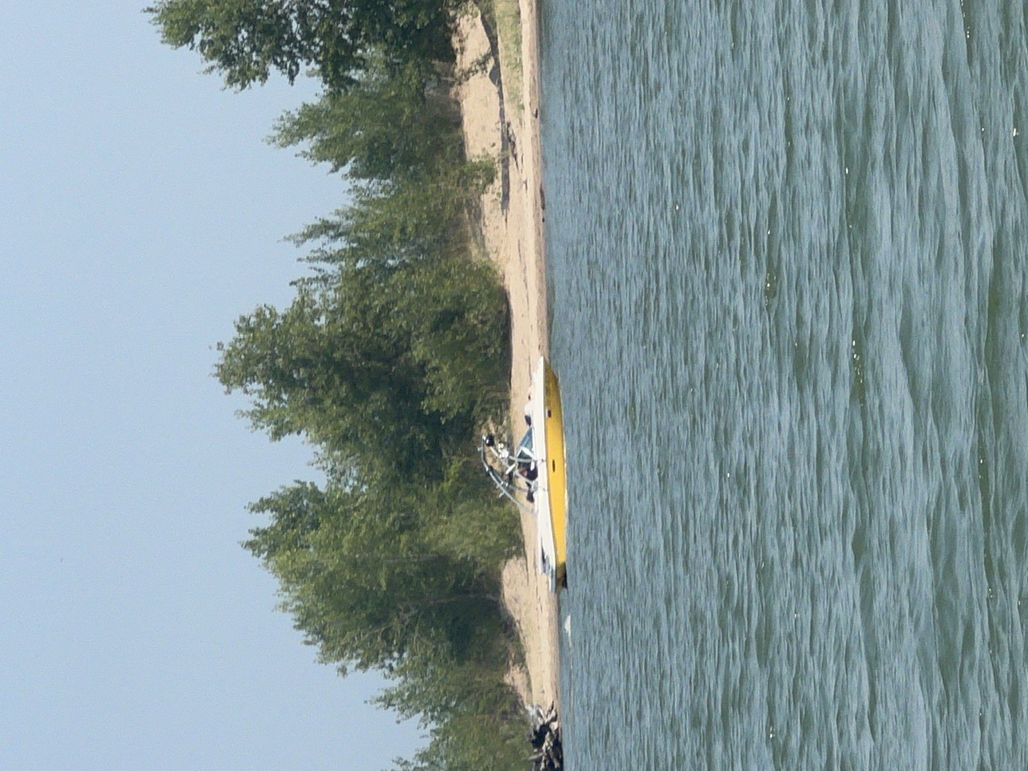 A yellow and white wakeboard boat is moving on a body of water. The boat is angled towards the shore, where trees and a sandy beach are visible. The water is choppy and the sky is clear and blue. There are no people clearly visible on the shore or in the water, however, there appears to be at least one person in the boat. The image appears to be taken from another boat or from the shore. There is no indication of the city of Borovoe, Russia in the image.