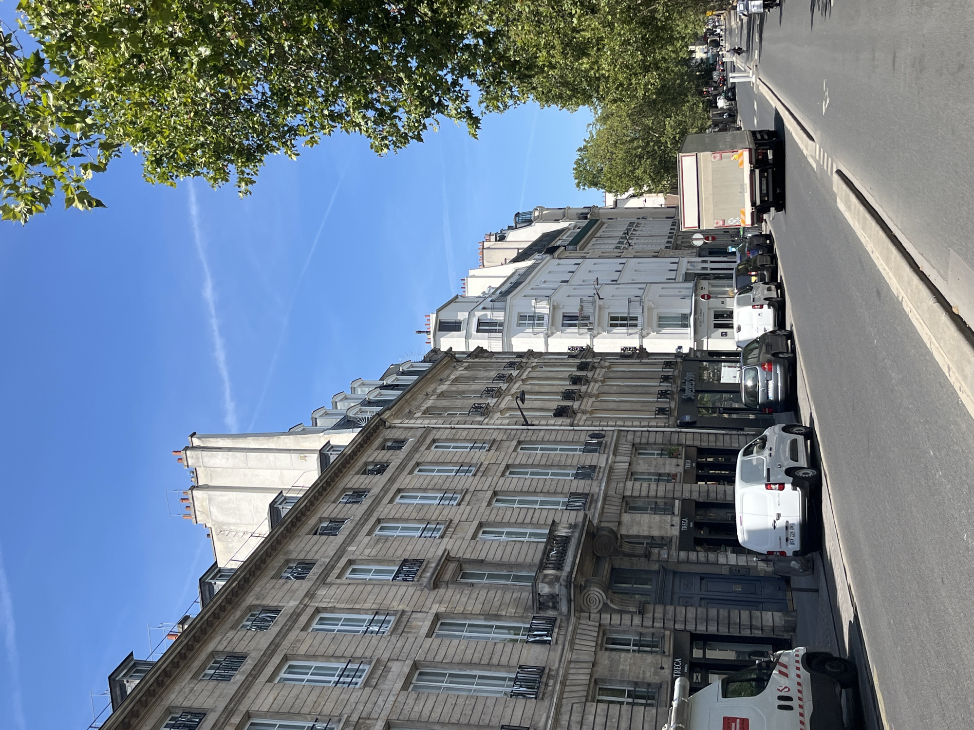 The image captures a street scene in Paris, France, on a clear day with a bright blue sky. The street is lined with tall, multi-story buildings, typical of Parisian architecture, with light-colored facades and many windows. On the left side, a row of older, stone buildings features ornate detailing, balconies with wrought-iron railings, and mansard roofs with dormer windows. Further down the street, the buildings transition to a lighter, white facade.
Several vehicles are parked or driving on the road. In the foreground on the left is a white van with red and white markings, followed by a white panel van, and then a dark-colored car. Further down the street, a large, beige truck is visible, its back end facing the camera. Trees with dense green foliage line the right side of the street, partially obscuring the buildings. The road is paved and appears to have a designated lane for traffic. A sidewalk runs alongside the buildings, and there are streetlights visible. In the distance, more buildings and trees can be seen, and the street seems to extend further into the city. There are a few people visible in the distance, appearing to walk or cycle. The overall impression is of a bustling urban environment on a sunny day.