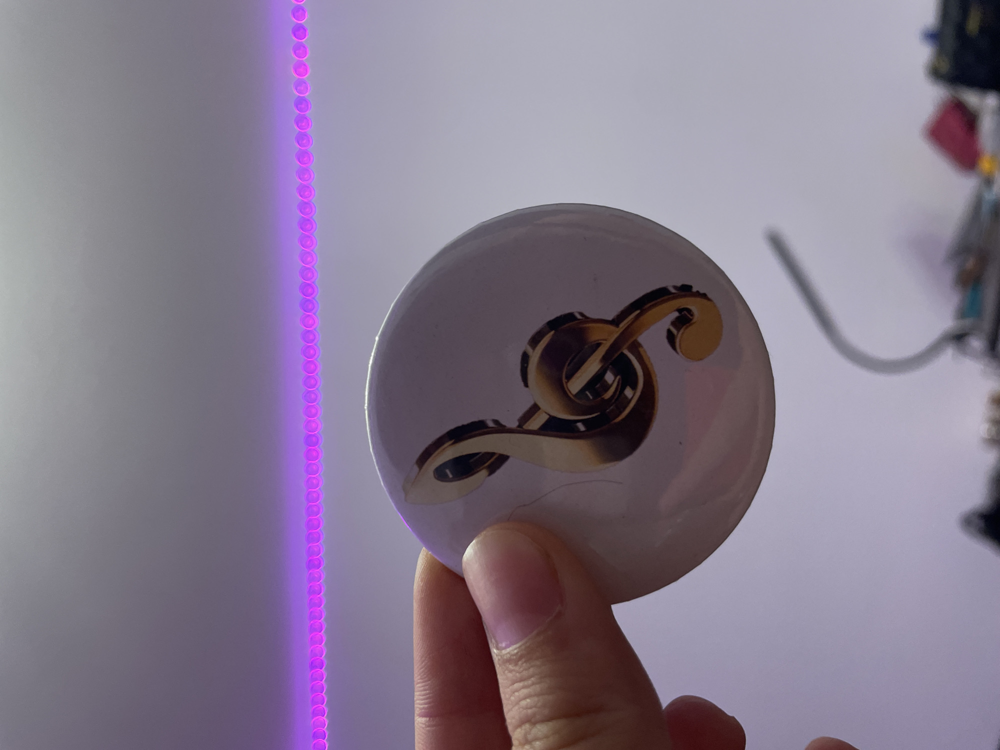 A person's hand is holding a round, white pin with a metallic gold treble clef symbol on it. The background is out of focus and appears to be a wall with a pink LED light strip running horizontally across the top. There are some blurred objects in the lower portion of the image. There is no visible indication of the location being Sinyavino, Russia.