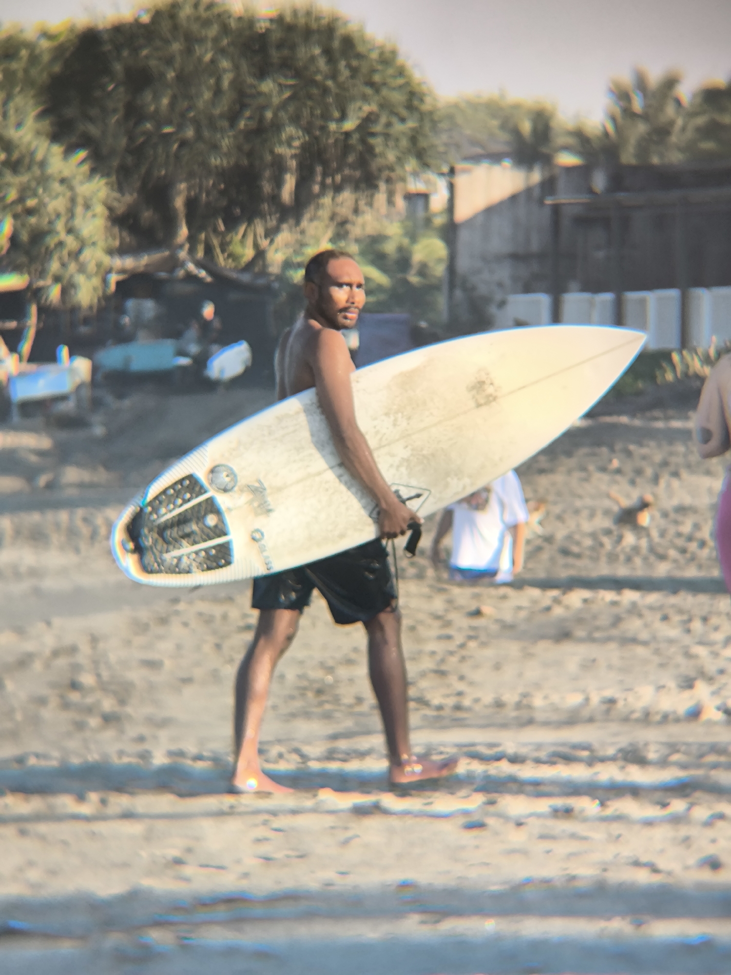 The image depicts a man on a beach in North Kuta, Indonesia. He is holding a white surfboard. The surfboard has black traction pads on the tail end and a small sticker on the nose. He is wearing dark-colored shorts. There is a partially visible person wearing a white shirt in the background. There are trees and buildings visible in the background. The sand appears wet near the bottom of the frame.