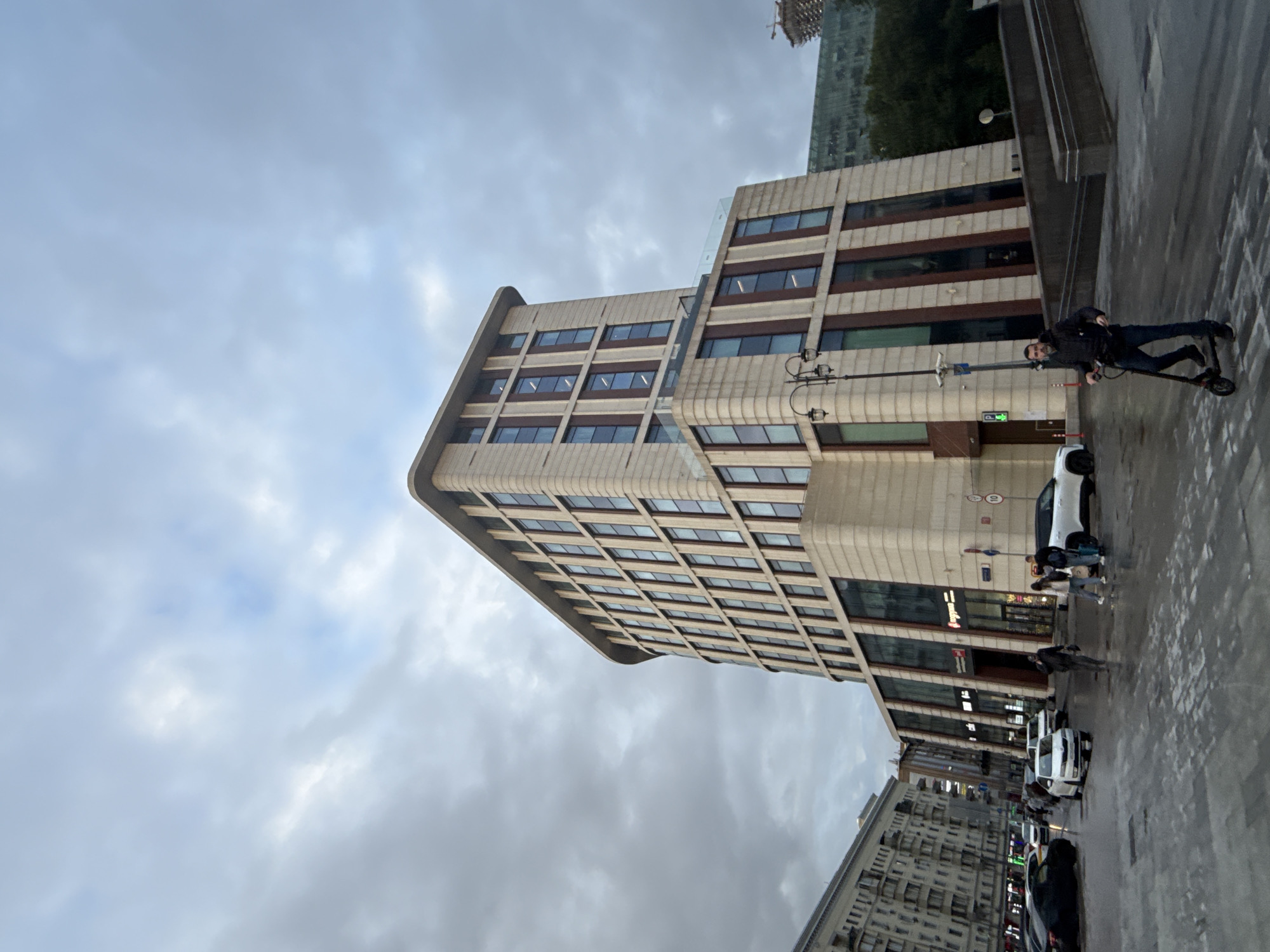 The image shows a low-angle view of a city street in Moscow, Russia, under a cloudy sky. A modern building with multiple floors and a curved facade dominates the scene. The building has numerous windows and brown horizontal accents. On the right side of the image, a man rides an electric scooter along the sidewalk. Several parked cars are visible along the street, and pedestrians are walking on the sidewalk near the building. In the background, there are other buildings, including one older, lower-rise building. The street surface appears wet, possibly from recent rain. Street signs are visible, though their content is not discernible.