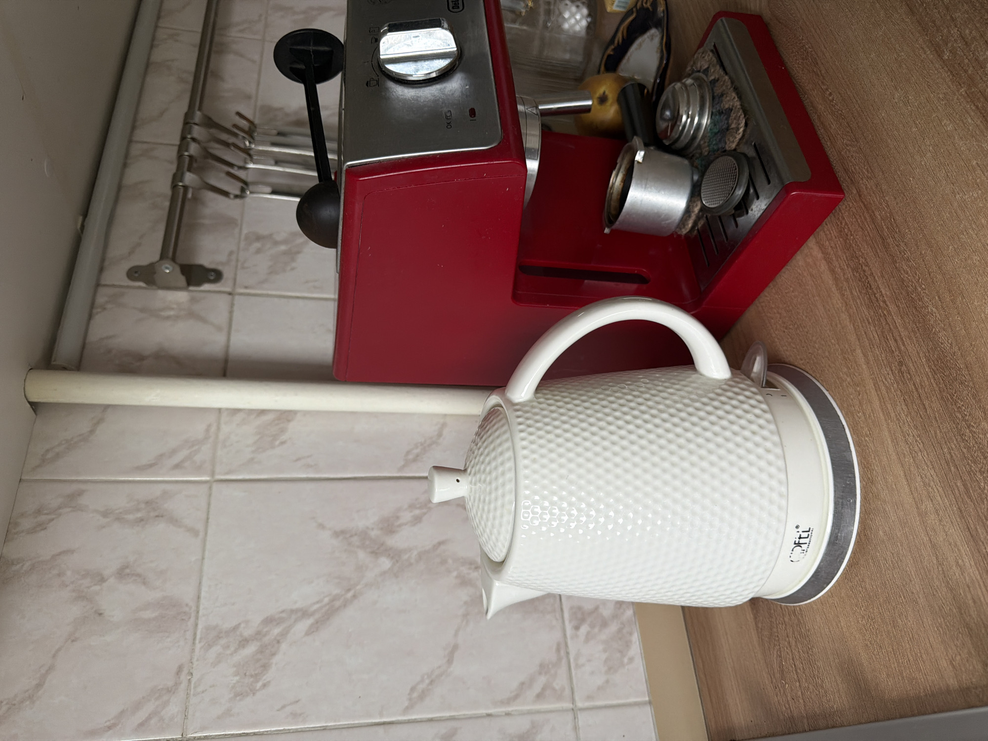 A red DeLonghi espresso machine and a white electric kettle are on a kitchen countertop. The espresso machine is on the left and the kettle is on the right. The background includes beige tile and a metal utensil rack. The location appears to be a domestic kitchen. The city and country information are not discernable from the image.