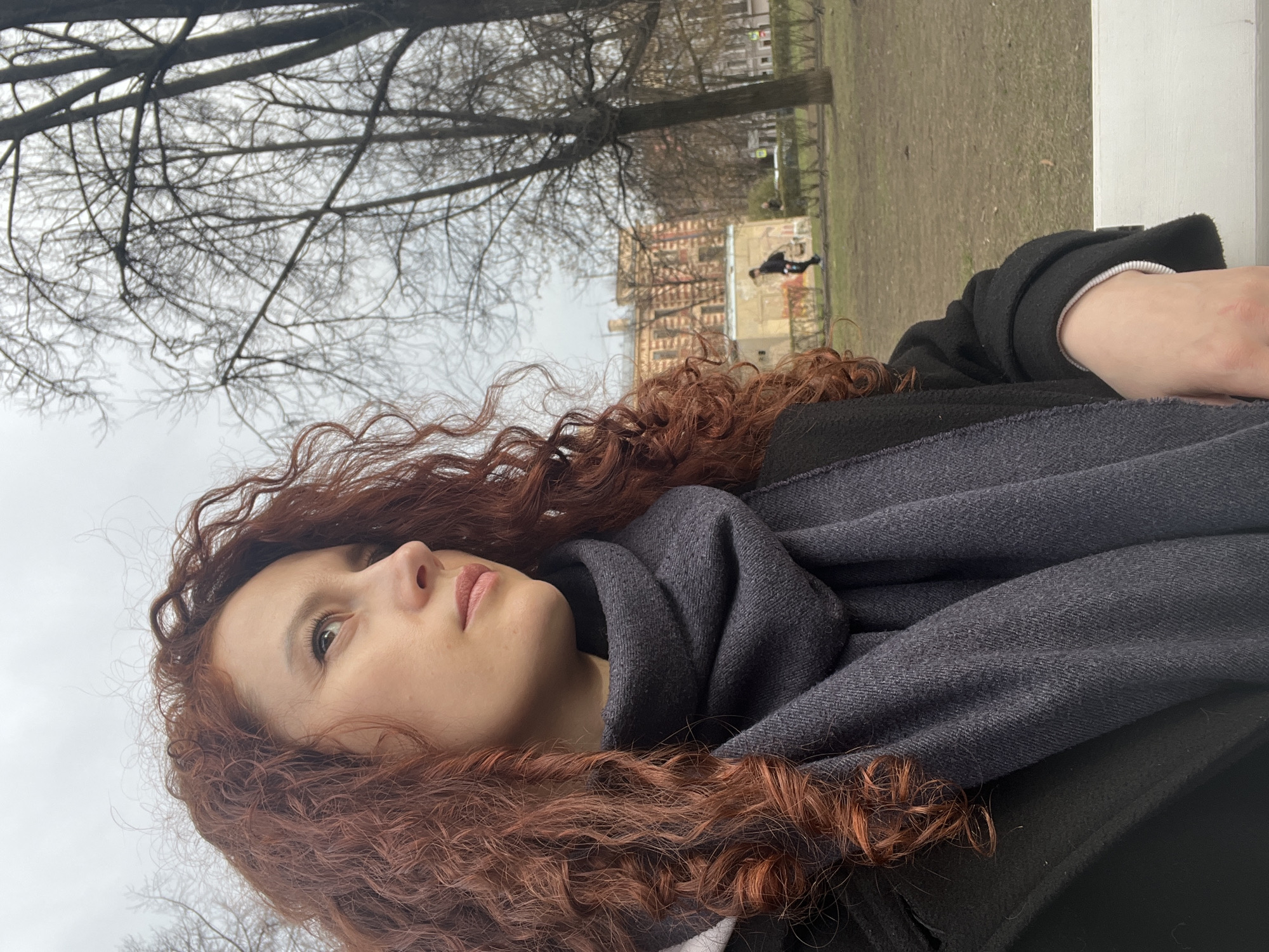 The image is an eye-level medium shot of a woman with long, curly red hair, wearing a dark coat and a gray scarf, looking upwards to the left. Her skin is light, and she has light-colored eyes and pink lips. The background is slightly out of focus, depicting a park landscape with bare trees and grass. A building is visible in the background, suggesting an urban environment.
Based on the architectural style of the building, specifically the brick and window design, and the overall park setting, the location is consistent with Saint Petersburg, Russia.
In the background, a person appears to be walking or standing on what seems to be a raised surface or structure. The lighting suggests an overcast day.
