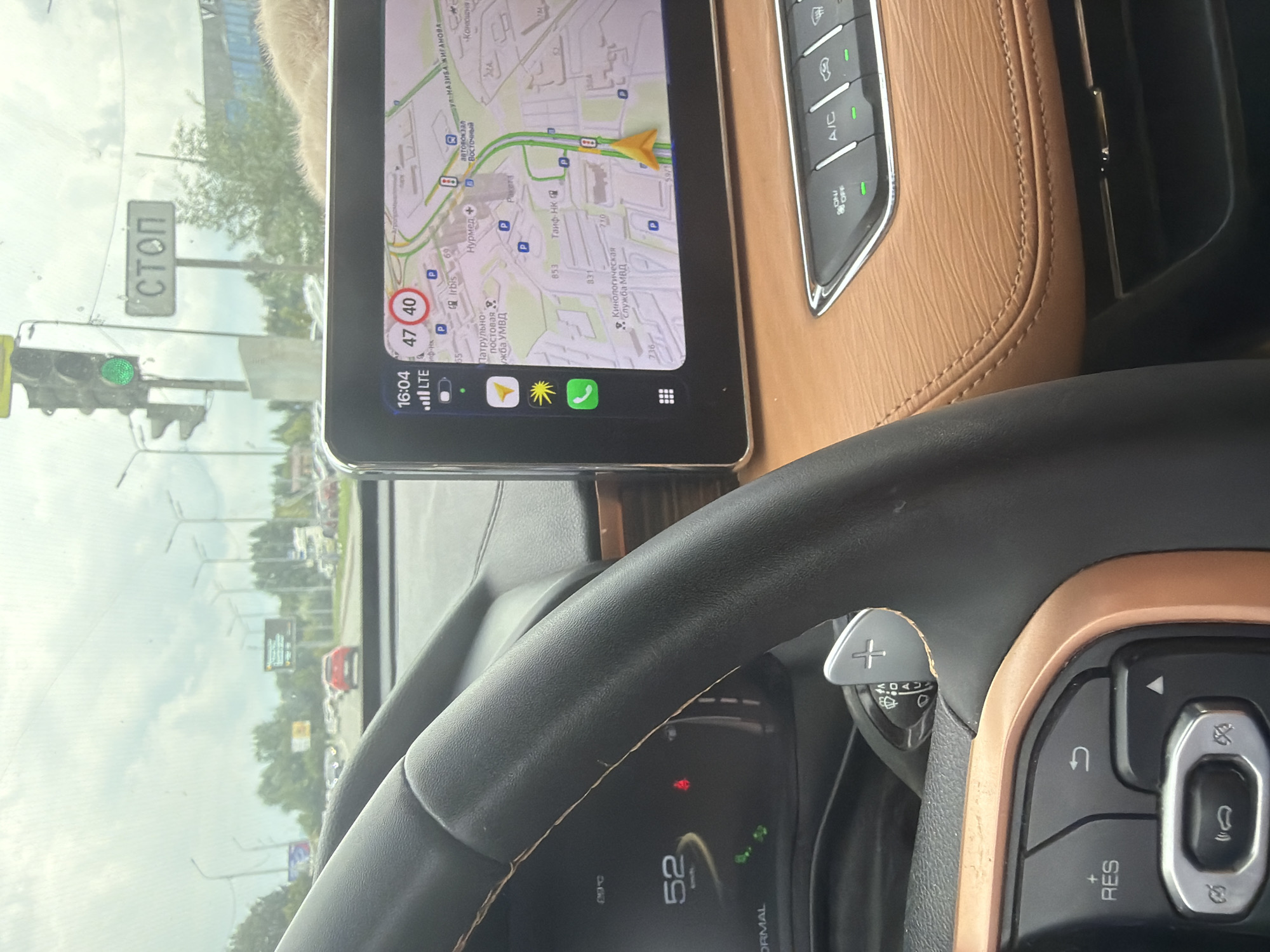 This is a photograph taken from the interior of a car, looking forward through the windshield. The car's dashboard is visible, including the steering wheel, instrument cluster, and a central infotainment screen.
The infotainment screen displays a navigation map, showing the location in Kazan, Russia, specifically on Prospekt Pobedy. The current time is 16:04, and the speed limit displayed is 40 km/h, with the car currently traveling at 47 km/h. Icons for a sun, phone, and home are visible.
The instrument cluster shows a speed of 52 km/h. The steering wheel is black leather with brown stitching. On the right side of the steering column are paddle shifters, marked with "+" and "-". The center console features climate control buttons, including A/C and "ON/OFF" indicators illuminated green.
Outside the car, the scene depicts a city street with multiple lanes of traffic, trees, streetlights, and traffic signals. A green traffic light is visible, and a "STOP" sign is mounted on a pole. Several vehicles are in traffic, including a red car in the distance. The sky is overcast with a cloudy, bright sky. The overall environment is a typical urban street scene on a cloudy day.