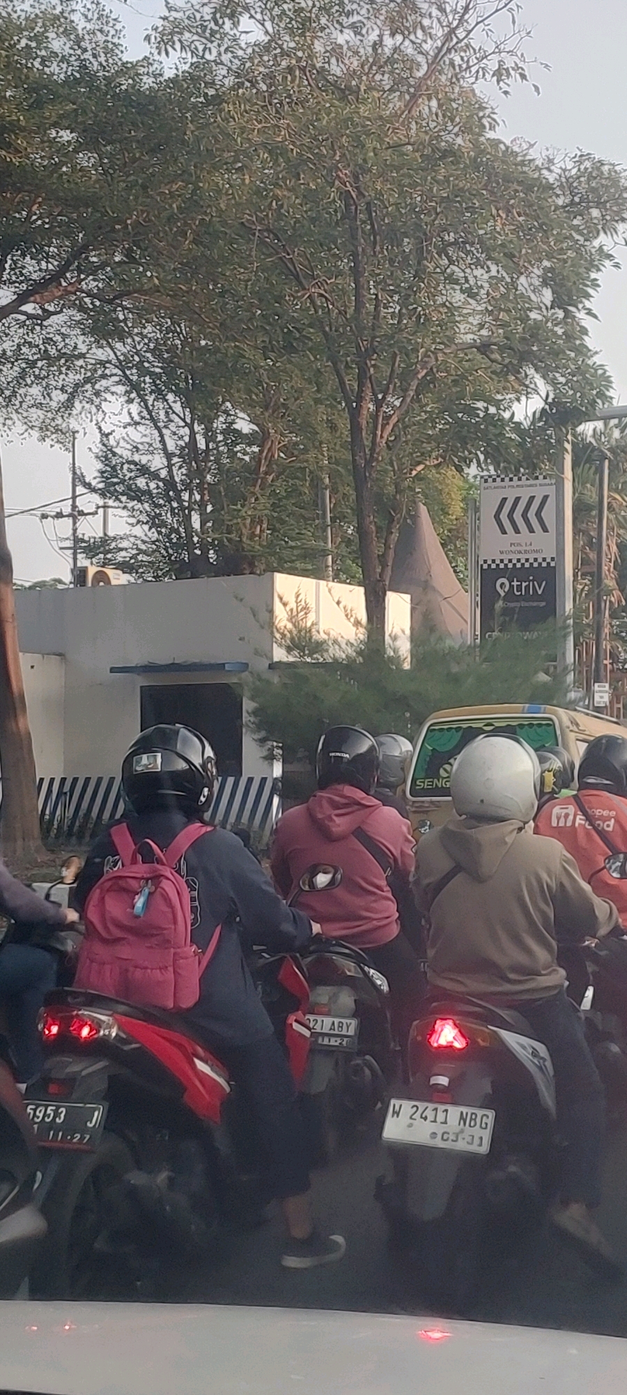 The image depicts a street scene in Surabaya, Indonesia, likely during daytime due to the lighting. In the foreground, several individuals are riding motorcycles in what appears to be moderate traffic.
On the left, a person wearing a black helmet and a dark jacket is riding a red motorcycle. They have a bright pink backpack on.
To the right of this motorcycle, another person in a dark helmet and a reddish-brown hooded sweatshirt is on a motorcycle. A dark strap is visible across their chest.
Further to the right, a person in a light brown hooded sweatshirt and a white helmet is riding a motorcycle. The back of an orange jacket with a "Food" logo is visible on another rider to the far right.
The motorcycles have visible license plates. The red motorcycle on the left has a plate reading "5953 J 1 11.27". The motorcycle in the center has a plate reading "21 ABY 11-20". The motorcycle on the right has a plate reading "W 2411 NBG CJ-31".
In the background, there is a light-colored building with a blue trim line. Above this building, trees with dense green foliage are prominent. A street sign with checkered patterns and the text "POS 1-4 WONOKROMO" and "triv Crypto Exchange" is visible on the right. A yellow vehicle, possibly a car or van, is partially visible behind the motorcycles. The overall environment suggests an urban setting with traffic and vegetation.