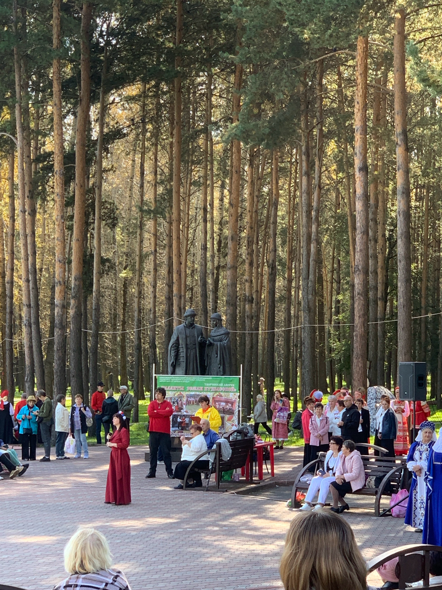 The image depicts an outdoor gathering in a park with tall pine trees. In the background, there is a statue of two figures and a banner with text in Russian. A group of people are present, many dressed in traditional folk costumes, suggesting a cultural event. Some individuals are standing and observing, while others are seated on benches. A woman in a long red dress stands center, appearing to address the crowd. The ground is paved with brickwork. The lighting suggests it is daytime, likely late afternoon given the shadows. The overall scene conveys a sense of community and celebration. The location is identified as a park in the city of Prokopyevsk, Russia.
