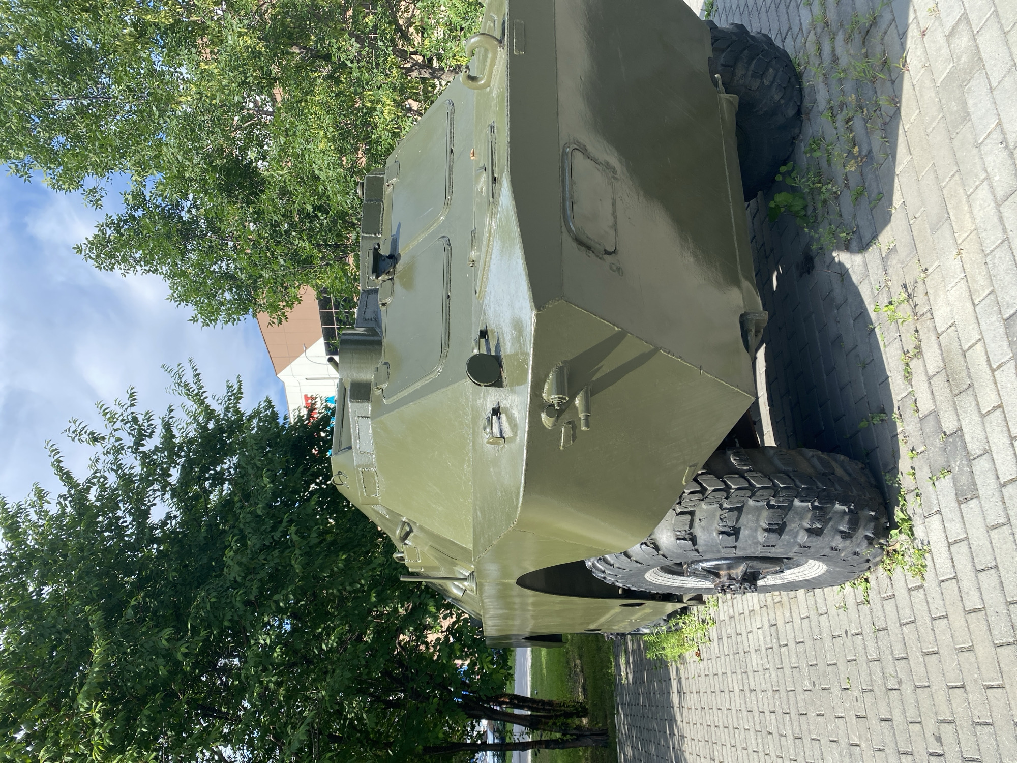A rear-view, medium-shot image displays a weathered, olive-drab armored personnel carrier parked on a brick-paved pathway. The vehicle, an apparent BTR-60 or similar model, shows signs of age with a dull, slightly scuffed paint finish. Its large, knobby tires are prominent, with one tire on the left side of the frame partially obscured by the vehicle's body. Green foliage from several trees forms a dense canopy overhead and frames the scene on the left and right, with a partially visible building in the background, appearing to be a light-colored structure with a reddish-brown section. The sky is a mix of blue with scattered white clouds. The setting is outdoors, likely in a public space or memorial park, given the paved walkway and manicured trees. No people are visible in the image. The overall impression is one of a static display, possibly part of a museum or monument, located in Novokuznetsk, Russia.