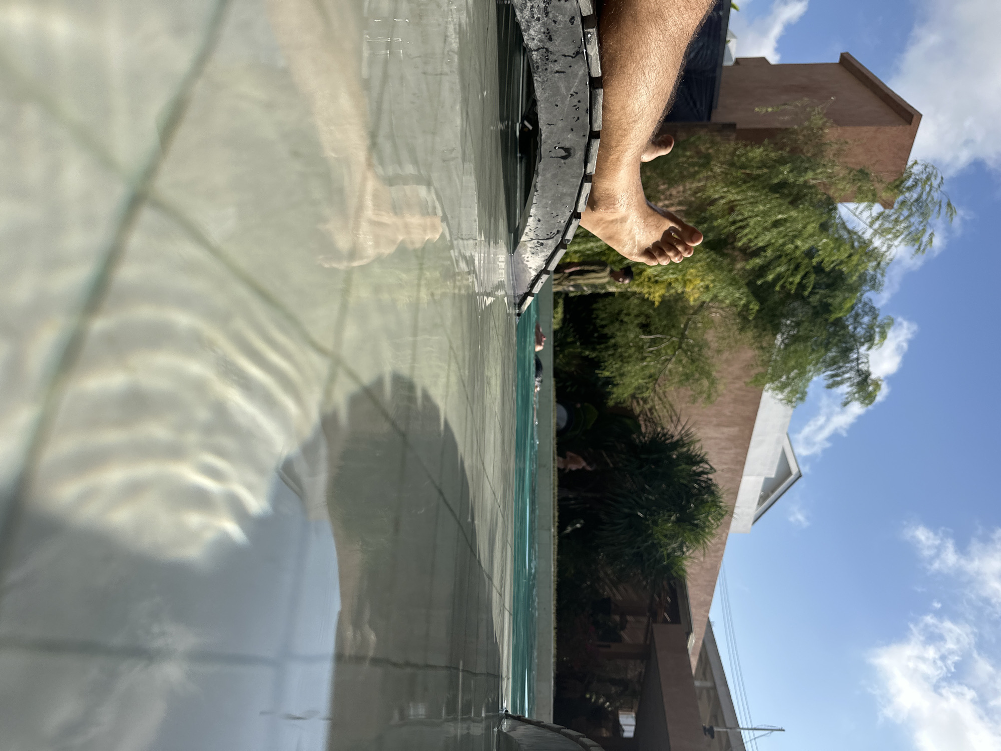 A first-person perspective shot shows a person's hairy legs and feet resting on a black lounge chair, with water reflecting the sunlight. The lounge chair is partially submerged in a swimming pool, where the tiled bottom is visible through the clear water. In the background, a building with a terracotta-colored facade and lush green trees are seen under a bright blue sky with scattered white clouds. Two other individuals are visible in the pool, one standing and another partially submerged. The scene suggests a relaxed atmosphere at a resort or private residence, likely in a warm climate. The location is identified as Kuta Selatan, Indonesia.