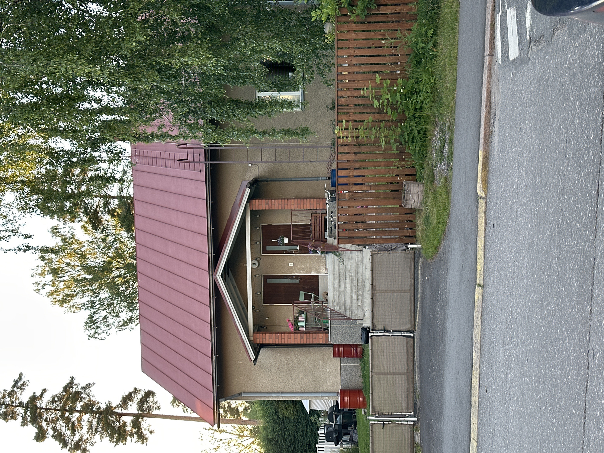 In an exterior, medium-shot, a two-story building with a red metal roof has a tan-colored stucco facade. Two identical, dark brown wooden doors with vertical paneling are visible on the ground floor, situated beneath a covered porch supported by brick columns. A set of concrete stairs leads up to the porch, flanked by metal railings. To the left of the entrance, a small metal table with two chairs and a potted plant sits on the porch. Red metal barrels are placed on the ground in front of the house, next to a white picket fence and grills. A wooden fence with vertical slats runs along the perimeter of the property, separating it from a paved area and a grassy lawn. Tall trees, including a pine tree, are visible behind the building. The scene is bathed in natural light, suggesting it is daytime.