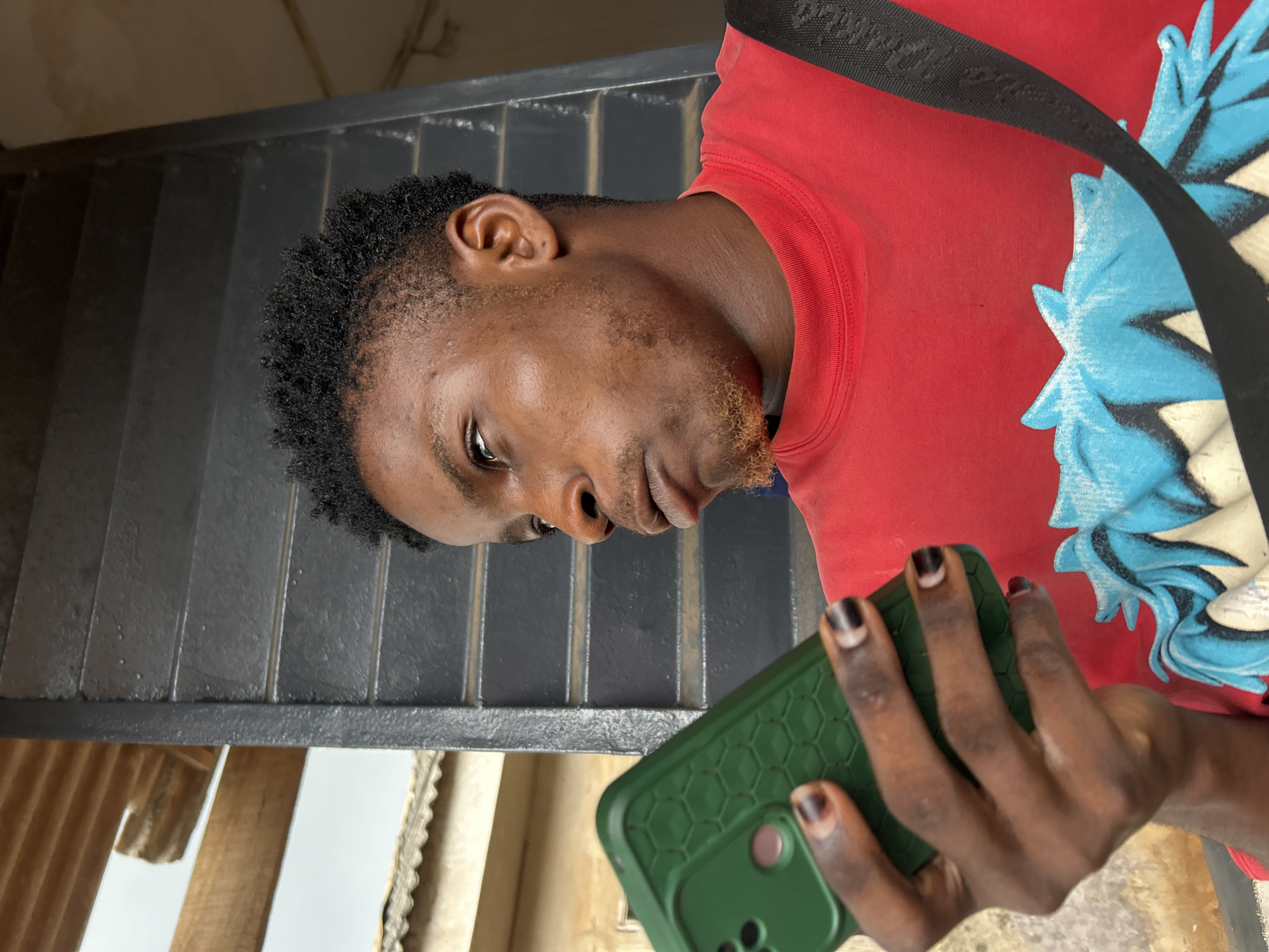 A man with short black hair and a reddish-brown beard is holding a green cell phone. He is wearing a red t-shirt with a blue and white design and a black strap across his body. He is standing in front of a black metal grate and wooden slats. The location is an unidentified area of Unknown, Unknown.
