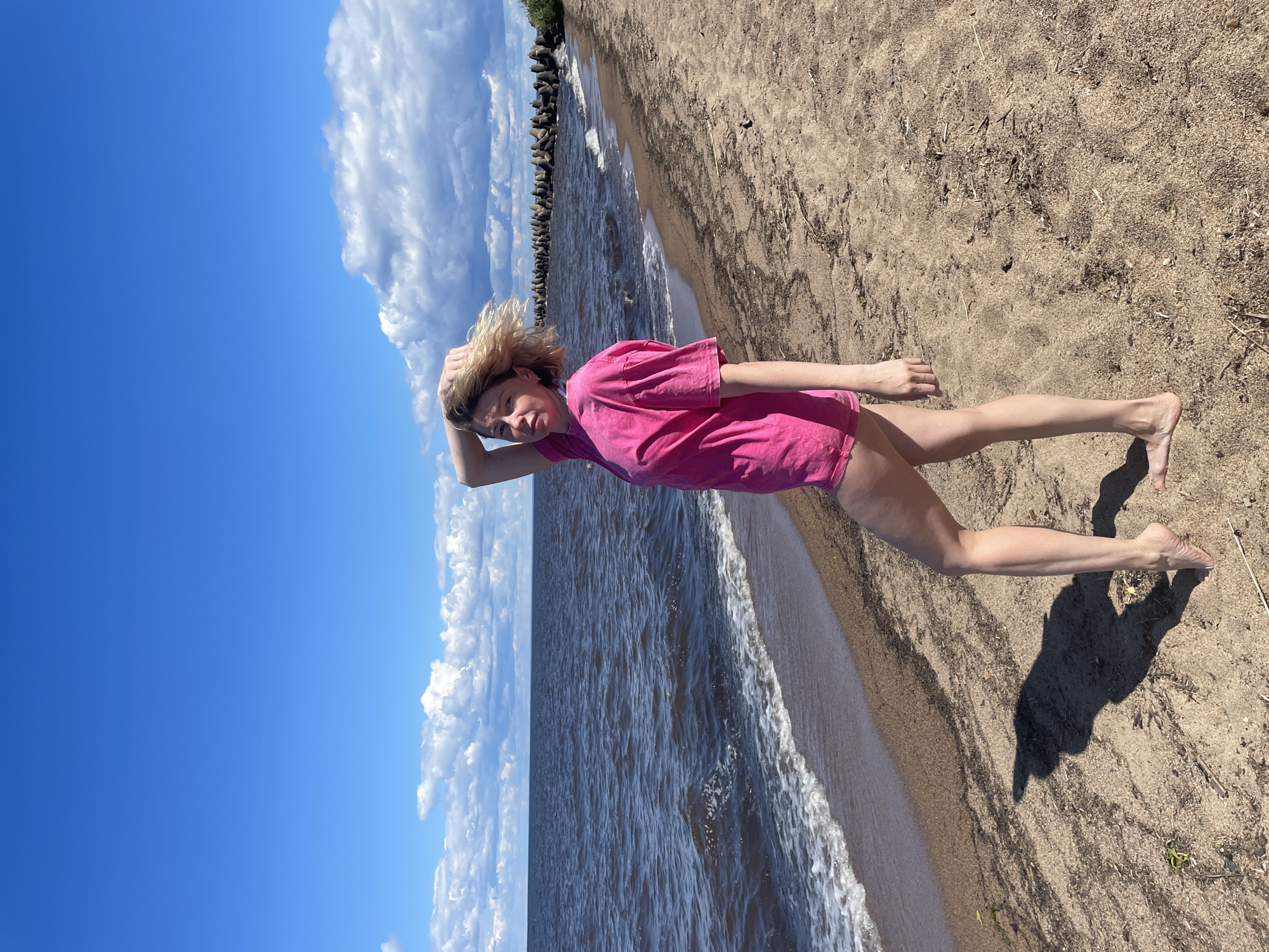 In the city of Belozersk, Russia, a blonde woman stands on a sandy beach. She is wearing a bright pink oversized t-shirt and appears to be posing with her hand in her hair. The woman is looking towards the camera with a serious expression. The beach is lined with dark water, and a line of concrete structures, possibly a breakwater, stretches along the coastline in the background. The sky is blue with scattered white clouds, and the sun is casting a shadow of the woman on the sand. The overall scene suggests a bright, possibly windy day by the water.