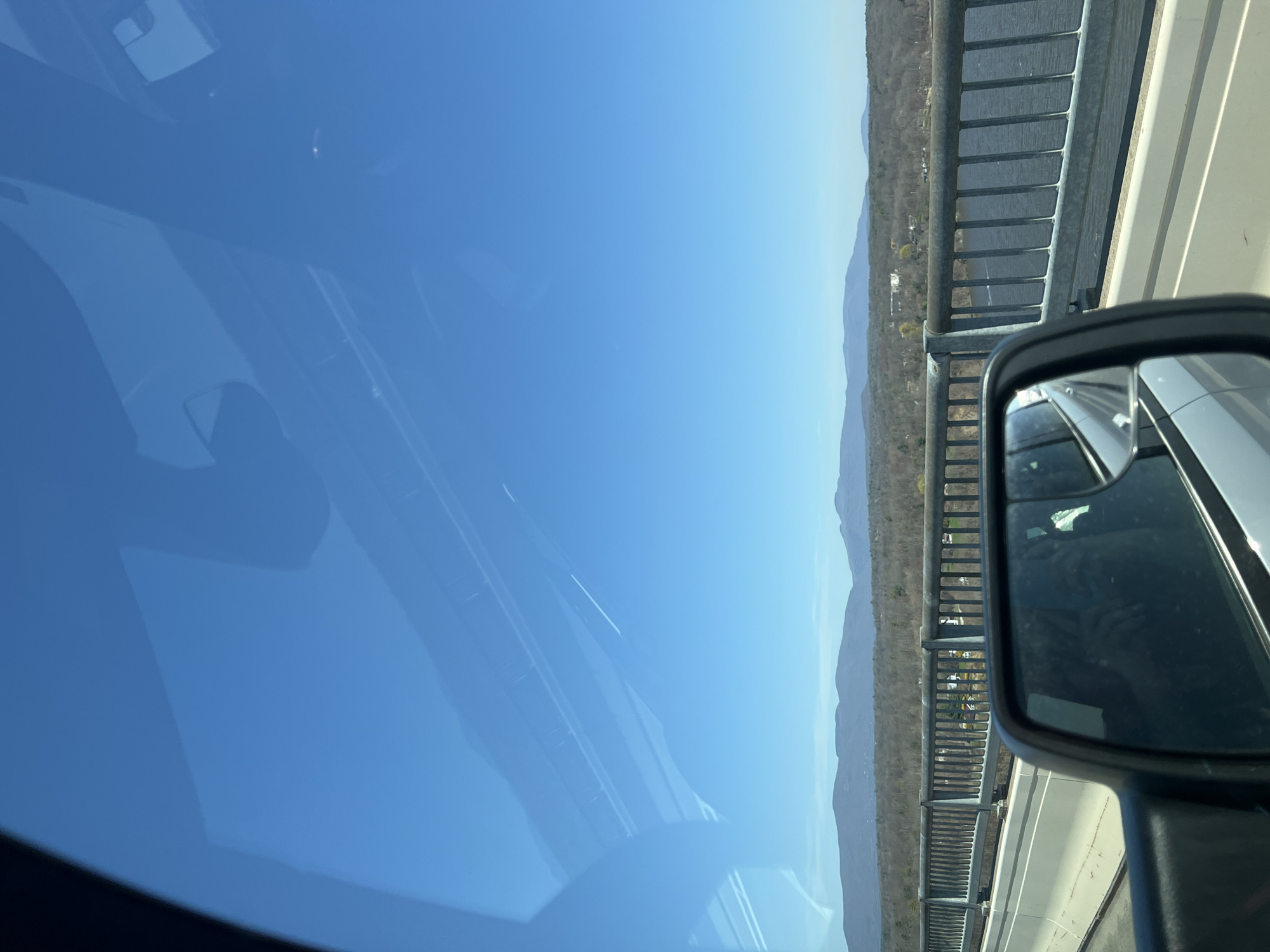 A car is traveling on the Kingston-Rhinecliff Bridge in Rhinebeck, New York, United States. The view through the windshield shows a clear blue sky. The bridge's railing is visible, as is the landscape beyond, including trees and distant mountains. The car's side mirror is also in view.