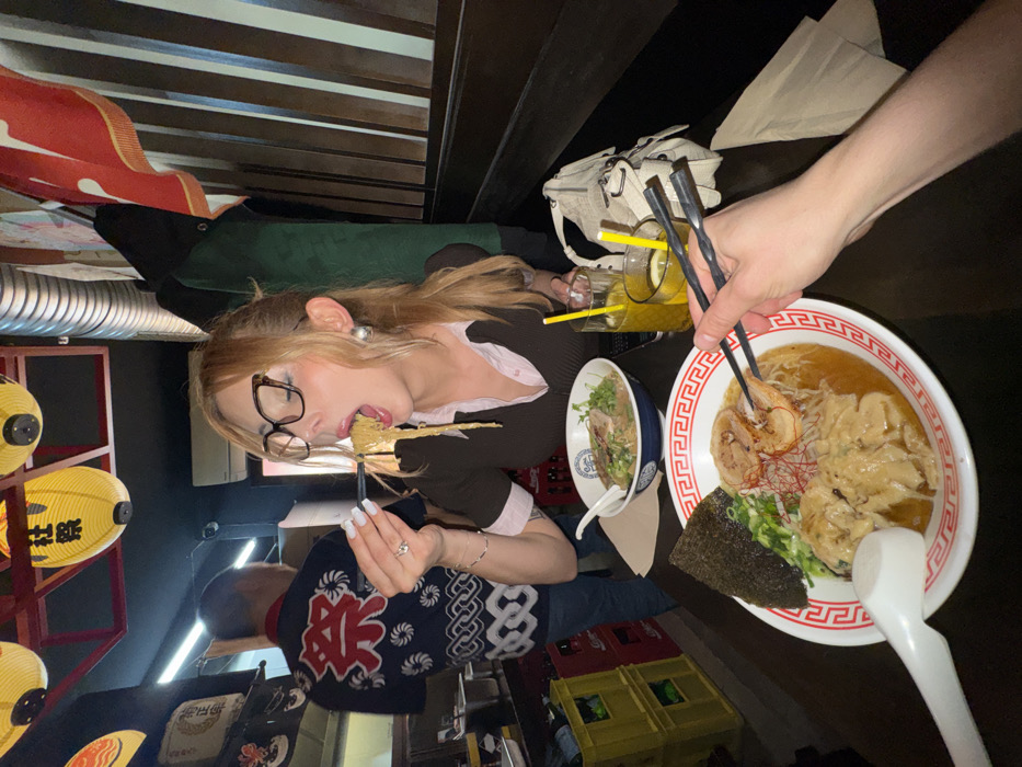 This image depicts an indoor dining scene at a Japanese ramen restaurant in Paris, France. A woman with light brown hair, wearing dark-rimmed glasses, a dark brown short-sleeved top, and a light pink collared shirt, is centrally positioned, actively consuming noodles with black chopsticks from a white bowl with a blue rim. Her mouth is open wide, holding the noodles. She has white nail polish, a ring on her left ring finger, and large, round, light-colored earrings.
In the foreground, a person's right arm extends into the frame, holding black twisted chopsticks over a white bowl of ramen. This bowl contains noodles, several slices of chashu (pork belly), green onions, bean sprouts, red chili threads, a sheet of nori (seaweed), and multiple dumplings. A white ceramic spoon rests in this bowl.
On the dark brown table surface, next to the woman, rests a white textured shoulder bag. Two clear glasses filled with a yellowish beverage and yellow straws are also present; one glass contains a lemon wedge.
In the background left, a man is partially visible from the back, wearing a dark blue shirt with a white traditional Japanese pattern and a large red character. Behind him, red plastic crates are stacked. Overhead, yellow Japanese paper lanterns hang from the dark ceiling, one prominently displaying the characters "社祭". The right side of the frame shows dark wooden paneling and a hanging red fabric banner.