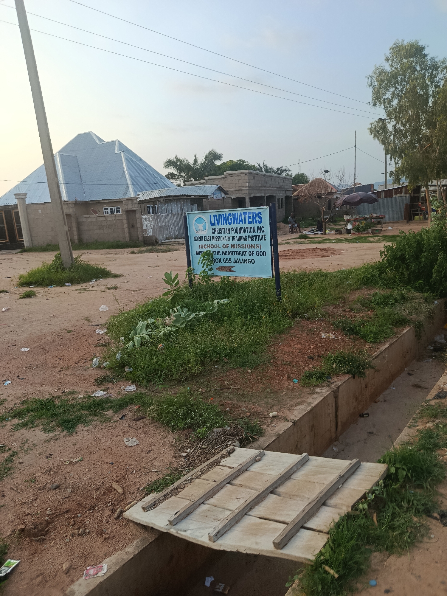 The image captures a street scene in Jalingo, Nigeria, likely during daylight hours given the lighting. In the foreground, a dirt road is visible with patches of grass and scattered debris. A concrete drainage ditch runs alongside the road, covered in places by a makeshift wooden bridge. A signpost stands prominently in the middle ground, displaying text that reads "LIVINGWATERS CHRISTIAN FOUNDATION INC. NORTH EAST MISSIONARY TRAINING INSTITUTE (SCHOOL OF MISSIONS) THE HEARTBEAT OF GOD P.O. BOX 695 JALINGO."
Behind the sign, several structures are present, including a building with a distinctive, angular, silver-colored metal roof, and other more modest dwellings with corrugated metal roofs. The environment appears somewhat rural or semi-urban, with a mix of developed and undeveloped areas. In the background, trees and utility poles are visible against a bright sky. No people are clearly identifiable in the image, though there are indistinct figures in the mid-ground near some of the structures. The overall impression is one of a functional, perhaps developing, community setting.
