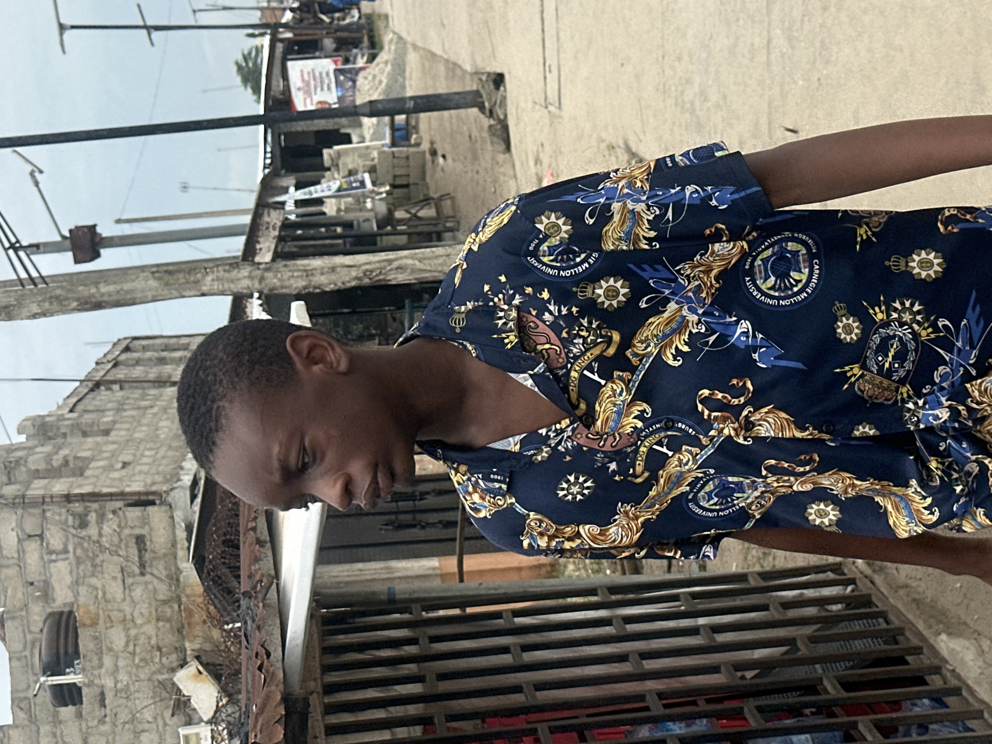 A young Black male stands facing left in the foreground, his head turned slightly towards the viewer. He is wearing a dark blue short-sleeved collared shirt adorned with a complex pattern of gold and blue motifs, including what appear to be crests and ornate scrolling designs. His skin tone is dark brown, and he has short, dark hair.
The background depicts a street scene in Badagry, Nigeria. To the left of the young male is a metal gate with vertical bars, behind which is a structure with a corrugated metal roof and rough-hewn stone walls. Further back, on the left side of the image, is a two-story building constructed of grey stone blocks, with a water tank visible on its roof.
The street itself is paved with concrete. Along the right side of the street, there are several open-fronted shops or market stalls, some with wooden beams and corrugated metal roofing. Signs and posters are visible on the stalls, including one with red and white text and another featuring a person's face. Utility poles with wires are scattered throughout the scene. In the distance, the sky is overcast. The overall environment suggests a bustling, possibly commercial, area in an urban setting.