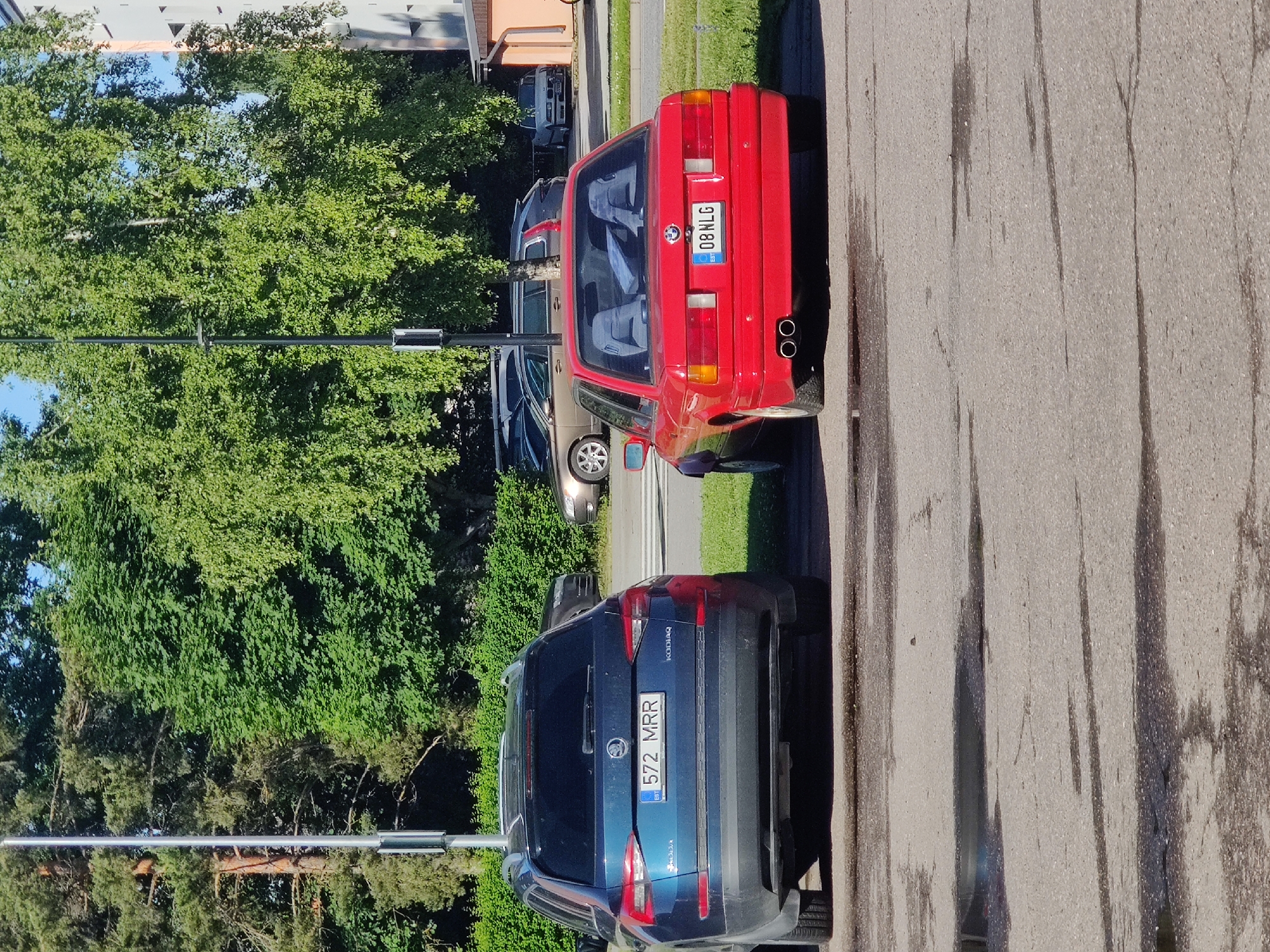 In Saue, Estonia, on a sunny day, two cars are parked parallel to each other in a paved lot. The car on the left is a dark blue Skoda Kodiaq, with its license plate displaying "572 MRR EST". The car on the right is a red BMW, identifiable by its license plate which reads "08 NLG EST". In the background, lush green trees form a dense canopy. A multi-story building with an orange facade is visible to the right of the BMW. The ground is asphalt, with scattered puddles reflecting the bright daylight. A metal pole, likely a lamp post, stands between the two cars and the trees.