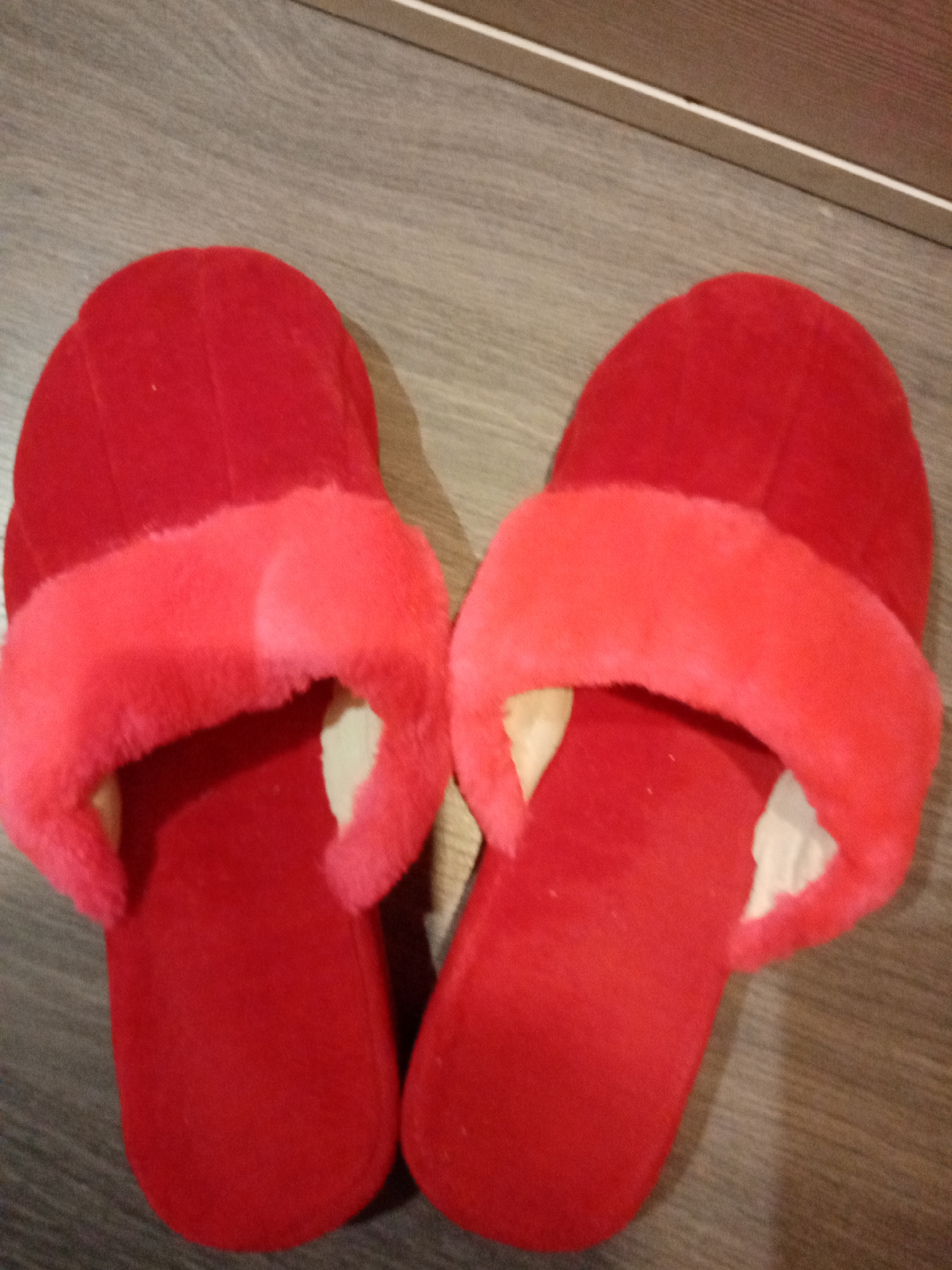 Two red slippers are on a wooden floor. The slippers have a slightly lighter red band across the top. The floor appears to be light grey in color. The upper right of the image shows the edge of a piece of furniture. No people, interactions, or city are discernible.