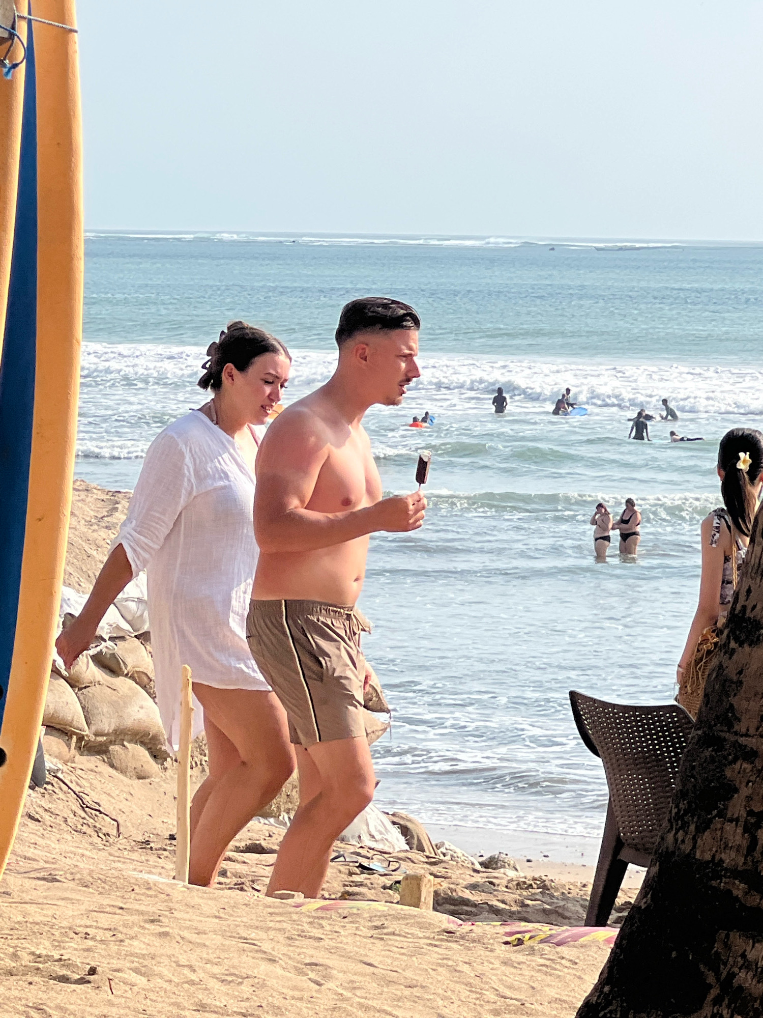 In Kuta, Indonesia, on a sunny day, a couple walks along a sandy beach adjacent to the ocean. The man, wearing brown swim trunks, holds an ice cream bar in his right hand and walks slightly ahead of the woman. The woman, dressed in a loose white tunic, is to his left. In the background, several people are in the ocean, some wading and others on surfboards. A surfboard with blue and yellow stripes stands vertically on the left side of the frame. The environment consists of sand, the sea with gentle waves, and a clear sky.