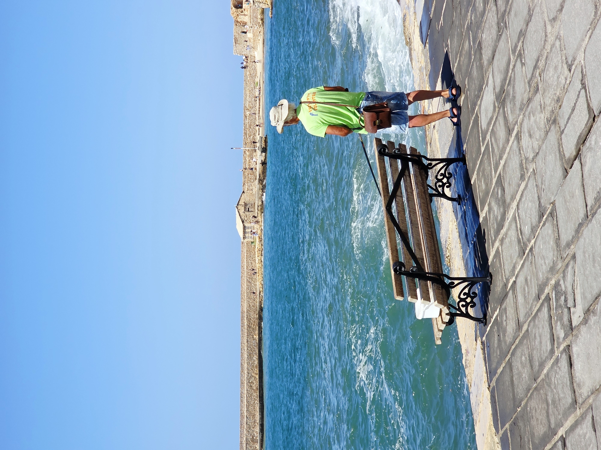 A man wearing a light-colored wide-brimmed hat, a bright green t-shirt, and denim shorts stands on a paved walkway next to a wooden bench. He is facing the sea, holding a fishing rod. A brown leather satchel is slung over his right shoulder. The sea is a vibrant turquoise with white-capped waves. In the background, a stone wall stretches along the coastline, with a few small buildings and people visible. The sky is a clear, bright blue. The scene is located at the harbor of Chania, Greece.