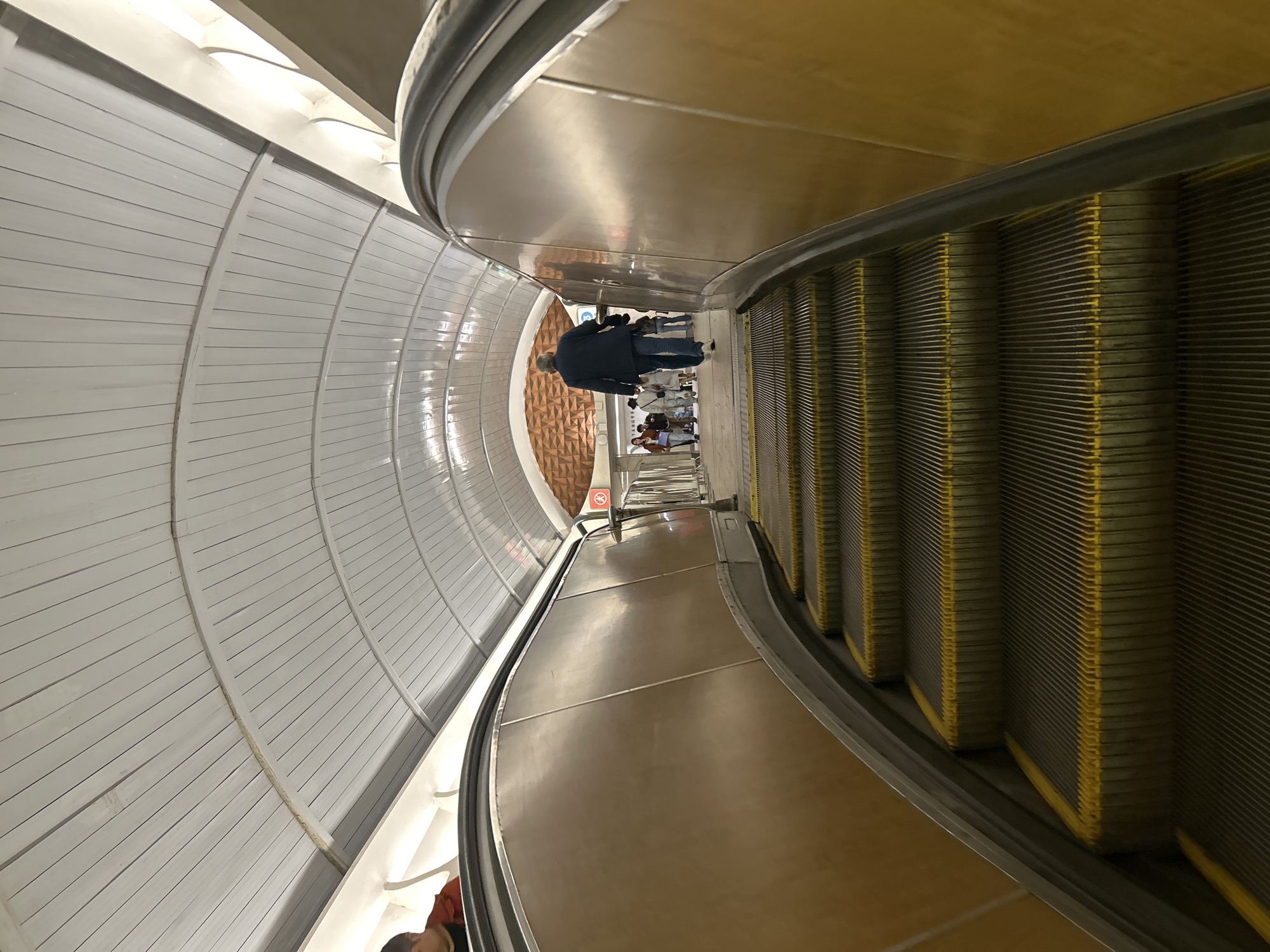 The image captures the perspective of an escalator journey downwards in what seems to be a subway station or a similar underground transport hub. Several individuals can be observed standing on the escalator. The walls are tiled, with a curved design featuring horizontal lines. The escalator's structure is metallic. The city of Unknown, Unknown can't be confirmed.