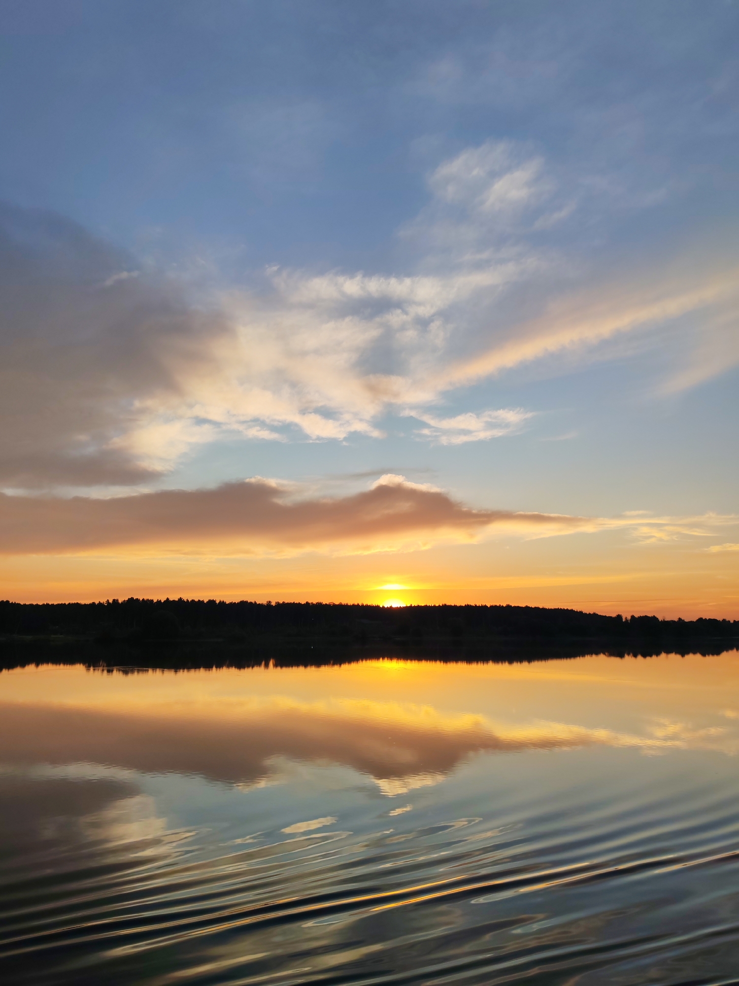 The image depicts a sunset over a body of water, likely a river or lake, near the village of Terekhovo, Russia. The sky is a gradient of blues, oranges, and yellows, with scattered clouds reflecting the warm hues of the setting sun. A dark, silhouetted treeline stretches across the horizon, marking the edge of the land. The water's surface is calm, with gentle ripples distorting the reflection of the sky and the treeline. The overall impression is one of natural beauty and tranquility during twilight.