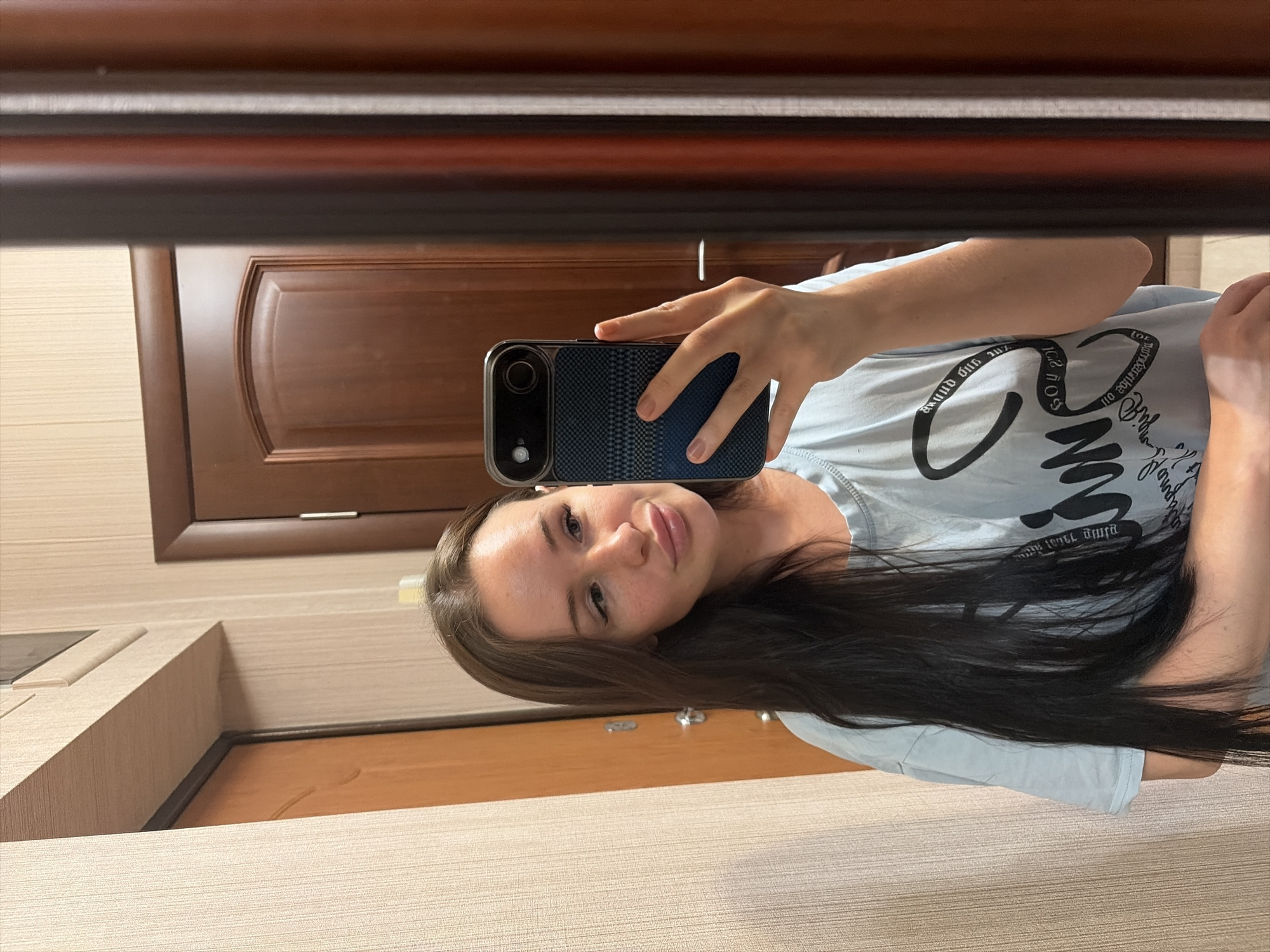 A young woman is captured in a mirror selfie, holding a dark-colored smartphone with a textured case in her right hand. She has long, dark brown hair and a neutral to slightly smiling expression, looking directly towards the camera reflected in the mirror. She is wearing a light blue t-shirt adorned with stylized, mirrored, and upside-down text, which is difficult to fully decipher but appears to include letters such as 'G', 'N', 'O', 'S', 'U', and 'L'.
The setting is indoors, featuring a background reflected in the mirror. Visible elements include two wooden doors with panel designs, one partially seen on the left and another more fully visible behind her. The walls are covered with a light-colored, horizontally striped wallpaper. A dark wooden frame or molding runs across the top of the image, suggesting it might be part of the mirror's frame or a piece of furniture above it. The lighting is bright, indicating it is likely daytime. The scene provides a glimpse into a casual, private space.